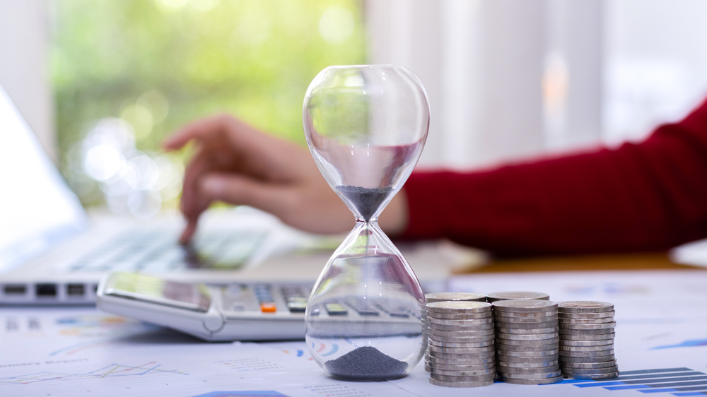 Münzen und Sanduhr vor einer Hand am Laptop