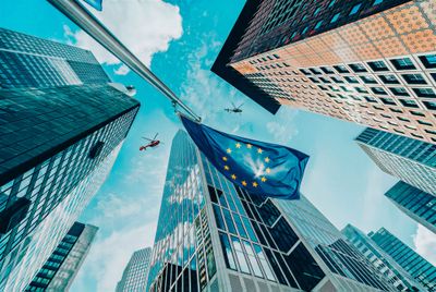 Blick von unten auf Hochhäuser mit EU-Flagge und zwei Helicopter darüber.