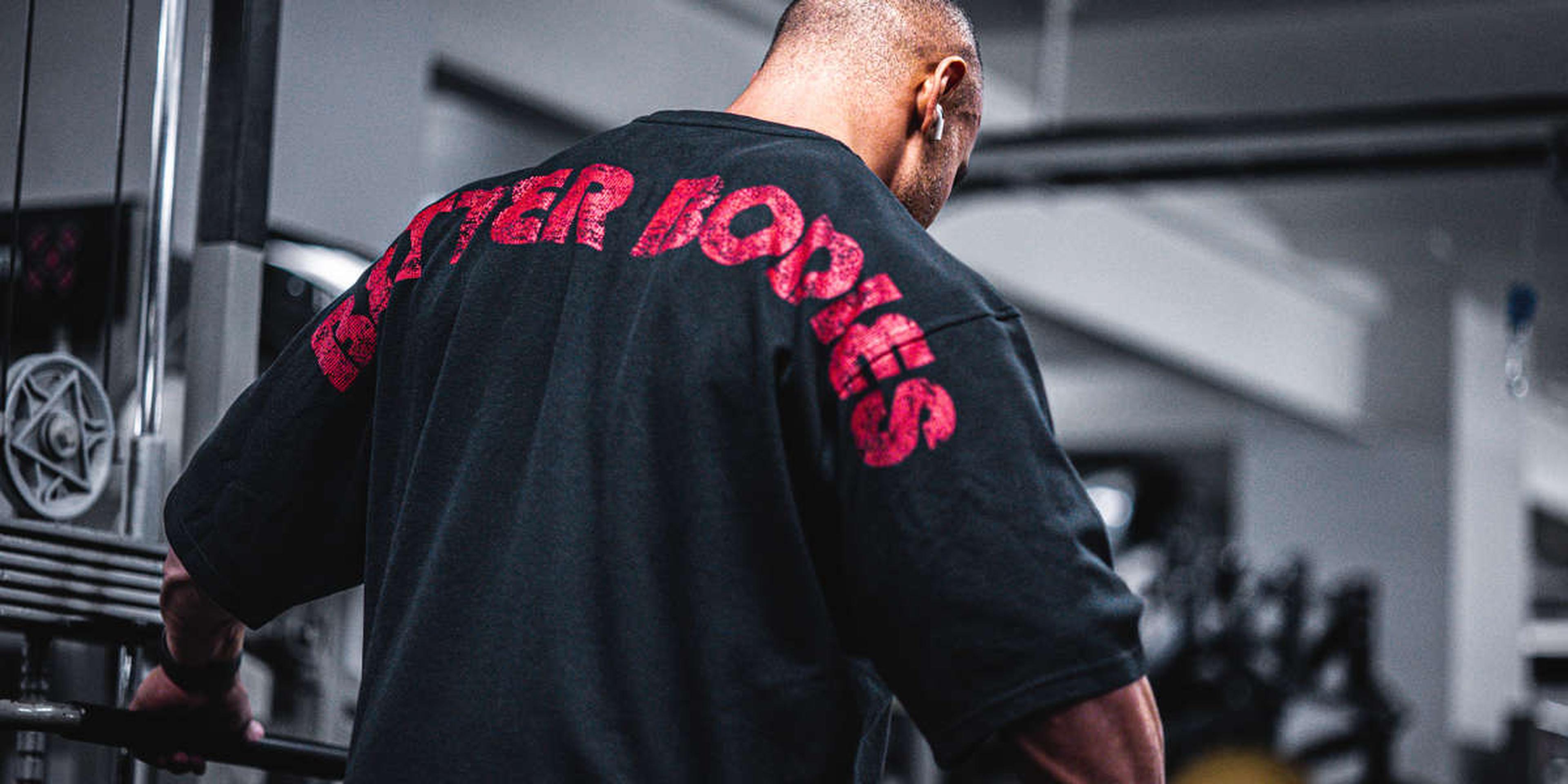 A person in a gym wearing a black shirt with "BETTER BODIES" in red letters, standing near exercise equipment, facing away.