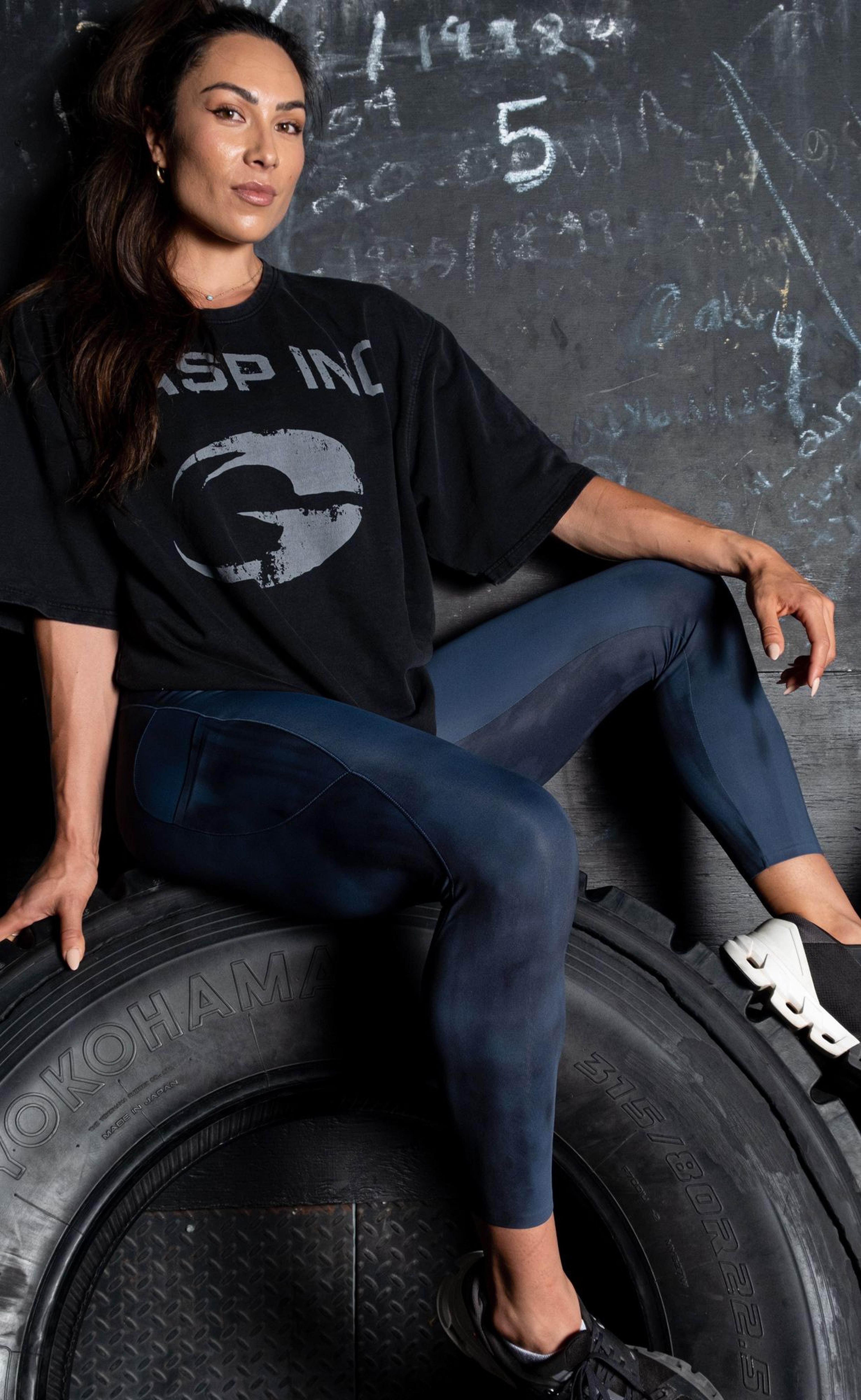 Woman in workout attire sits on a large tire against a chalkboard wall, wearing a black T-shirt and blue leggings.