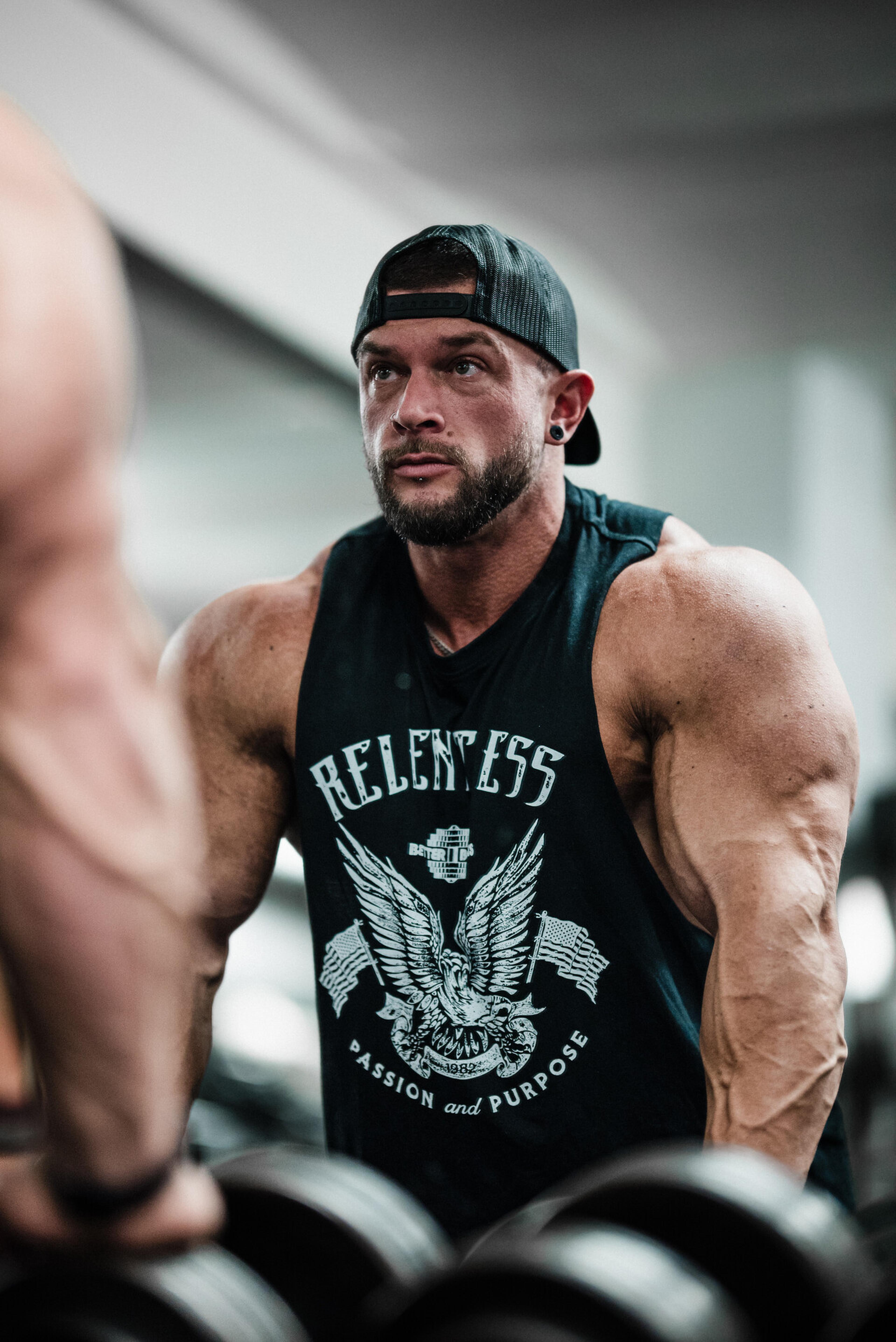 Man in a gym wearing a "Relentless" tank top and cap, intensely focused while lifting weights, showcasing muscular arms.