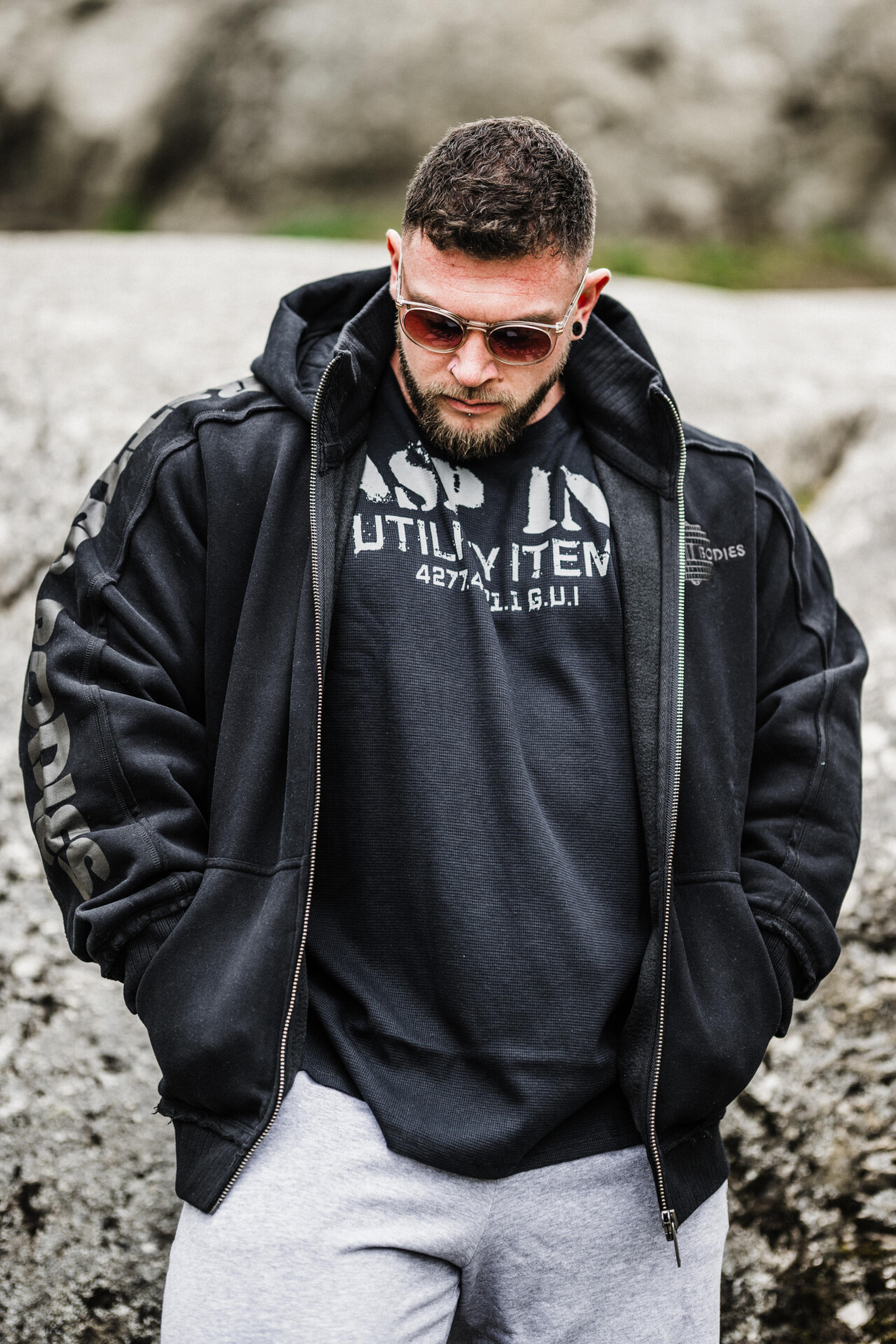 Man in sunglasses and a black hoodie stands outdoors, looking down. He wears gray sweatpants, with rocky terrain in the background.