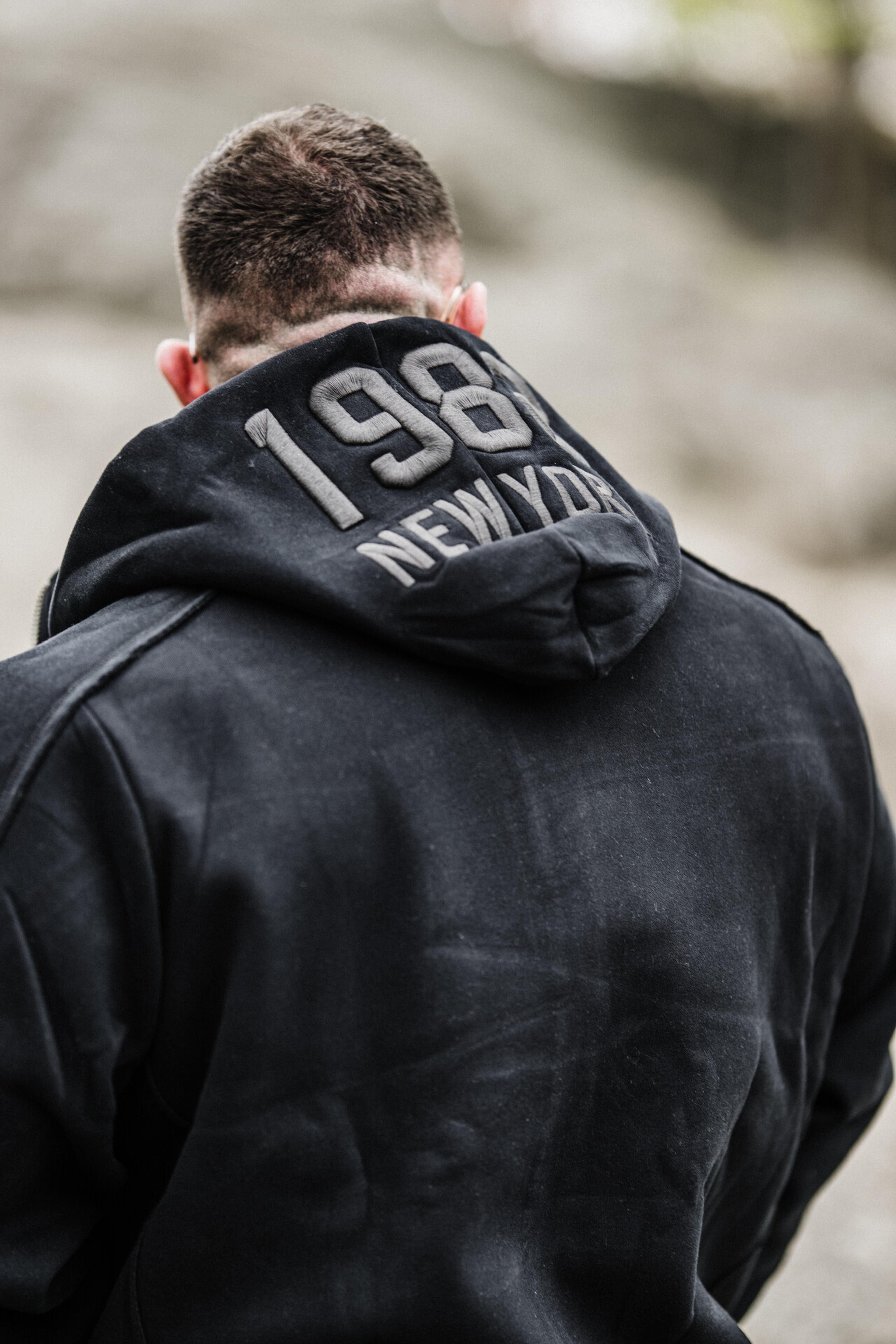 Person wearing a black hoodie with "1984 New York" printed on the back, standing outdoors with a blurred background.