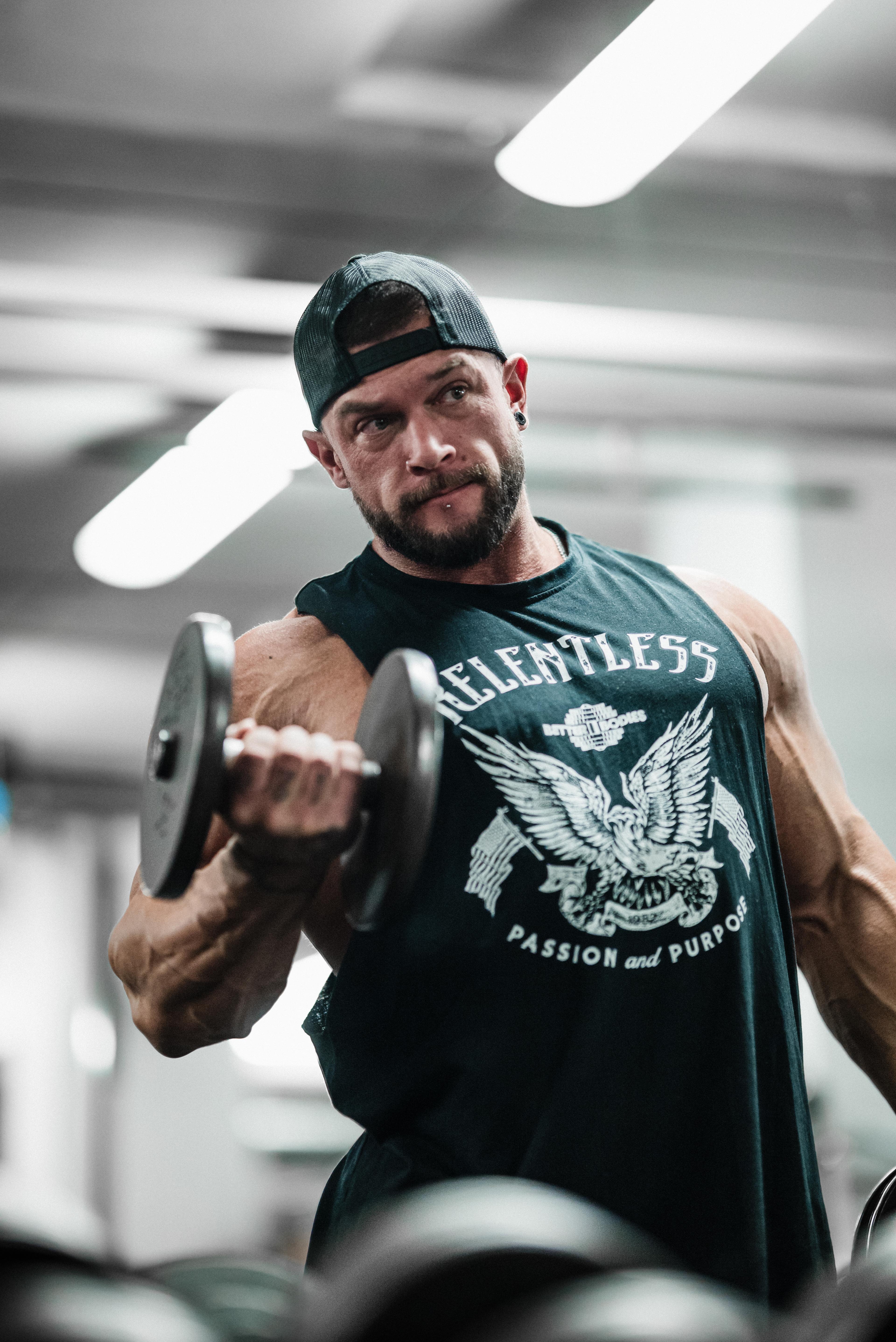 A muscular man in a gym performs a bicep curl with a dumbbell, wearing a black cap and sleeveless shirt with "Relentless Passion and Purpose."