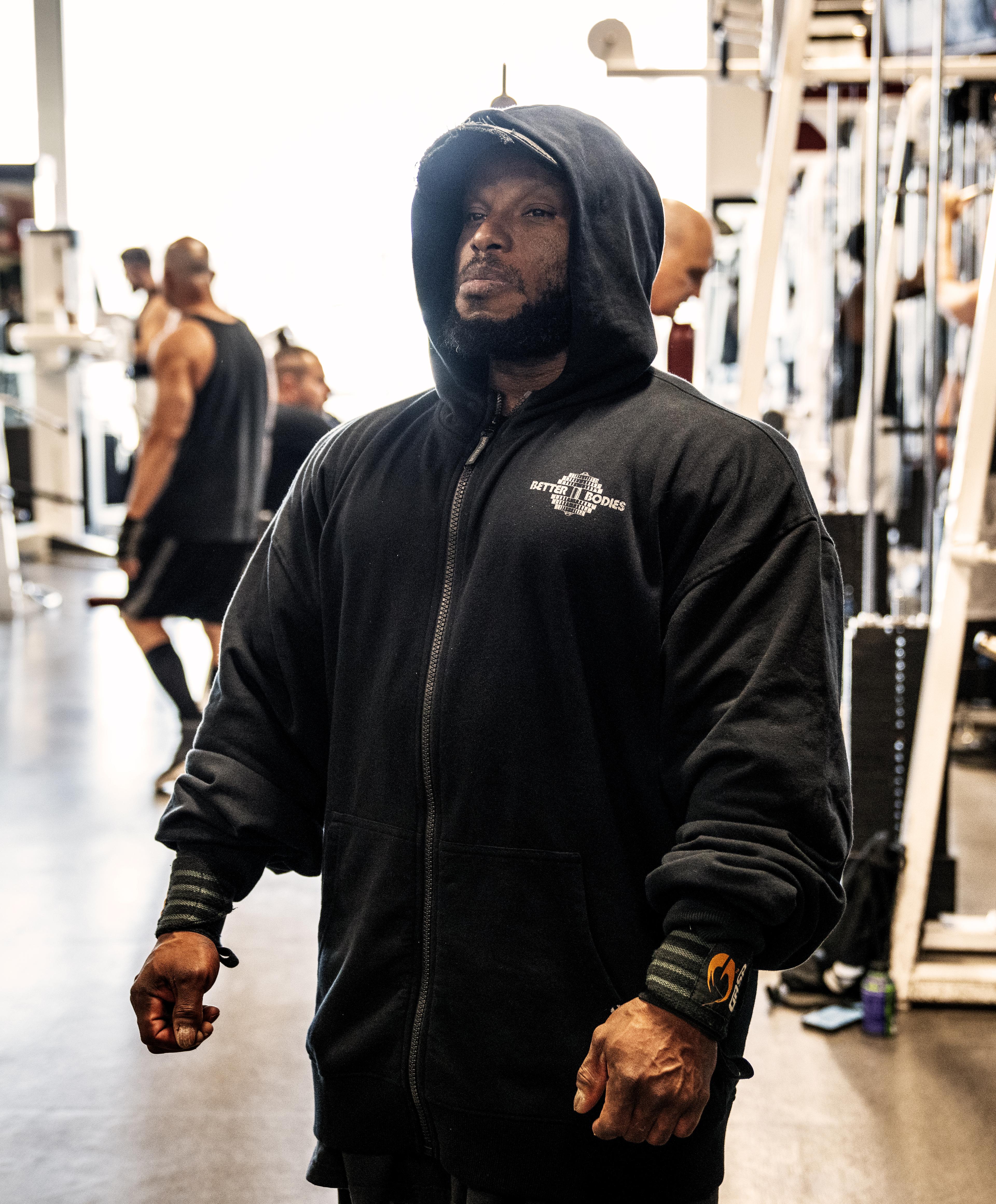 Person in a gym wearing a black hoodie, standing with a focused expression. Gym equipment and other individuals are visible in the background.