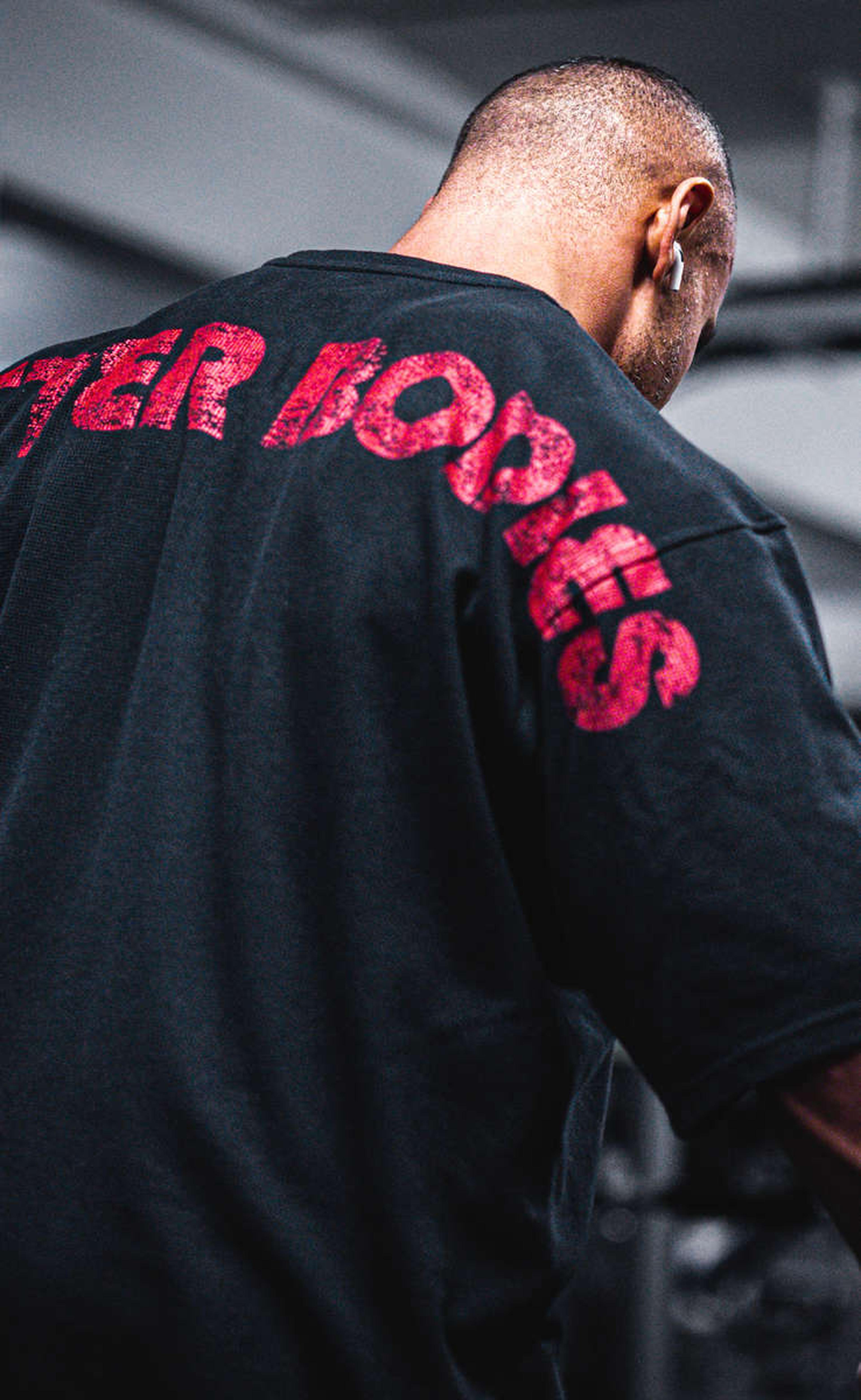 Man in a gym wearing a black shirt with "BETTER BODIES" in red letters on the back, standing against a blurred background.