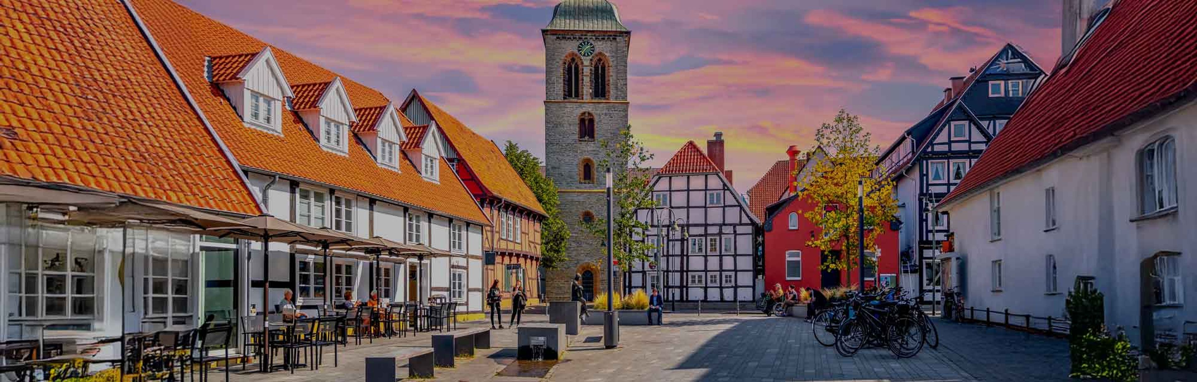 Historic European town square with cobblestone streets, half-timbered buildings, red-tiled roofs, a central church tower, outdoor seating, and a colorful sunset sky.