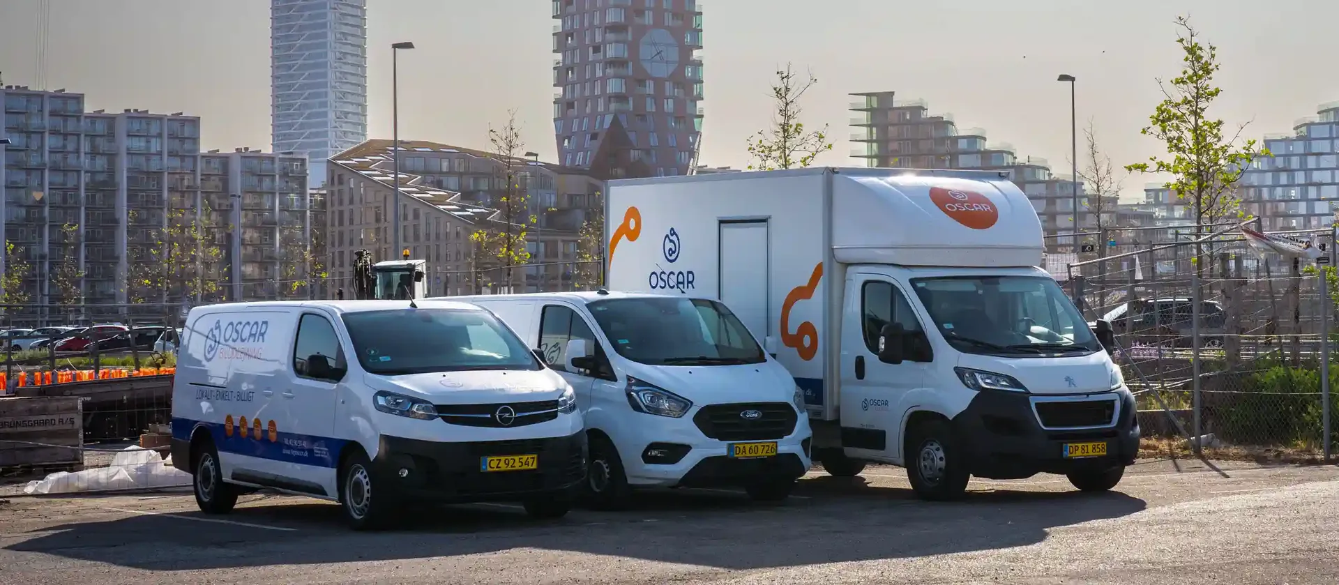 Two white Oscar Car Rental vans and an Oscar Car Rental box truck parked side by side in an urban lot, with modern high-rise buildings and young trees in the background.