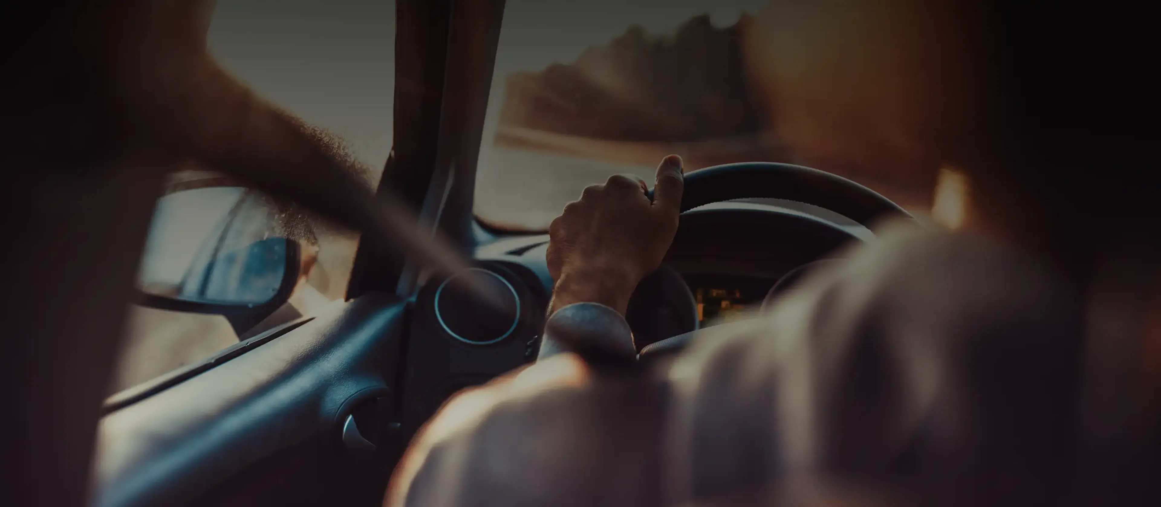 Close-up view from the back seat of a person driving a Oscar Car Rental car, with one hand on the steering wheel and warm sunlight illuminating the interior