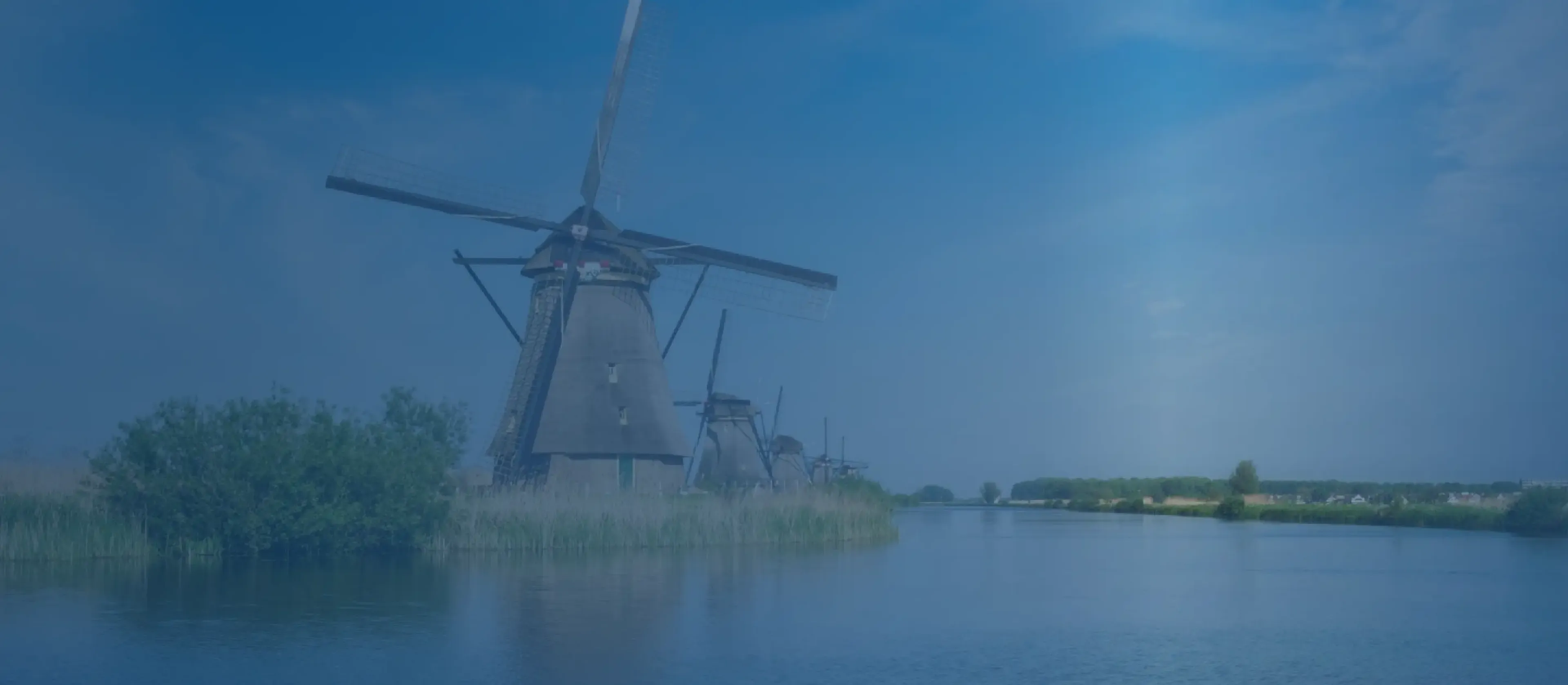 Five traditional Dutch windmills along a calm canal under a blue sky, reflections on the water and grassy banks.