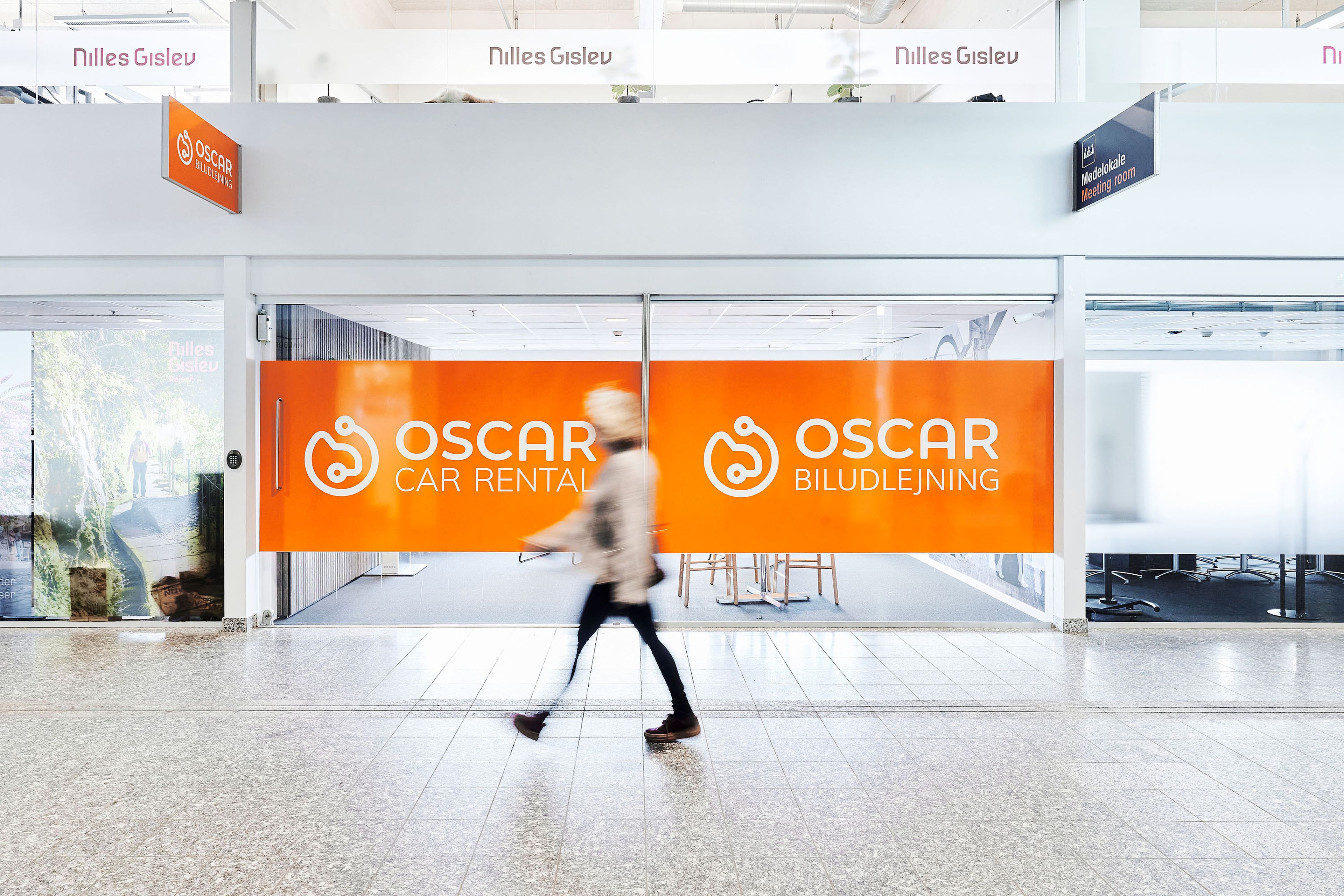 Man kneeling beside orange "Oscar Car Rental" banner outside a small rental office with several parked cars.