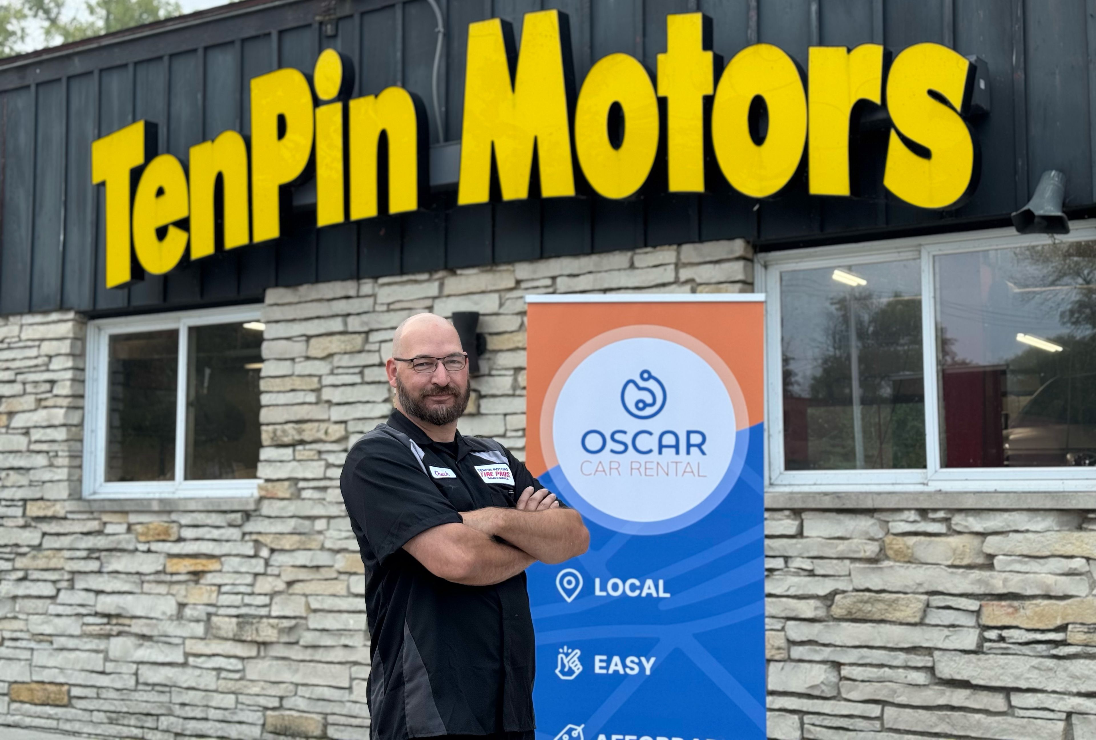 Man in black shirt standing with arms crossed by a blue "Oscar Car Rental" banner in front of TenPin Motors storefront