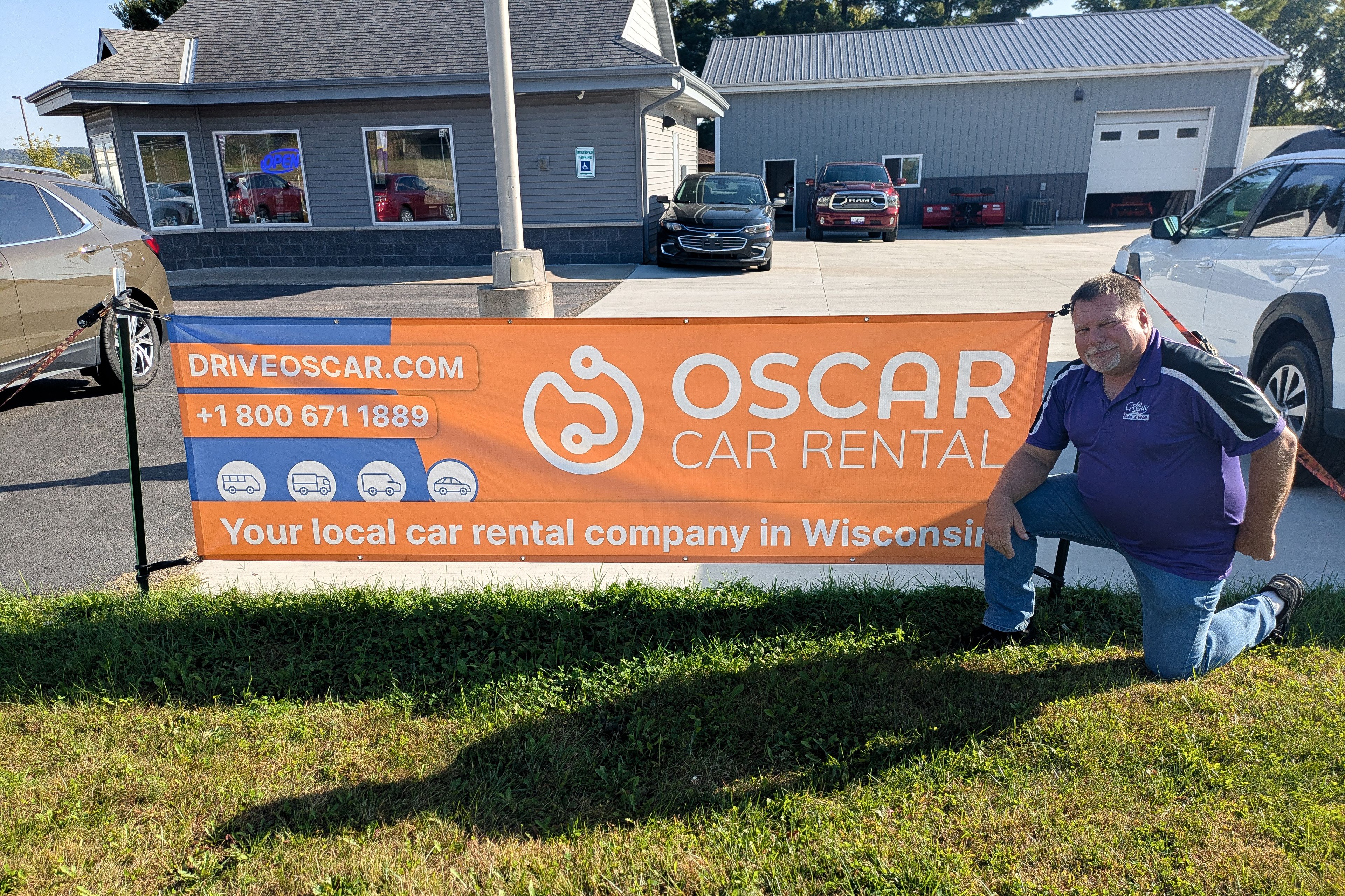 Man kneeling beside orange "Oscar Car Rental" banner outside a small rental office with several parked cars.