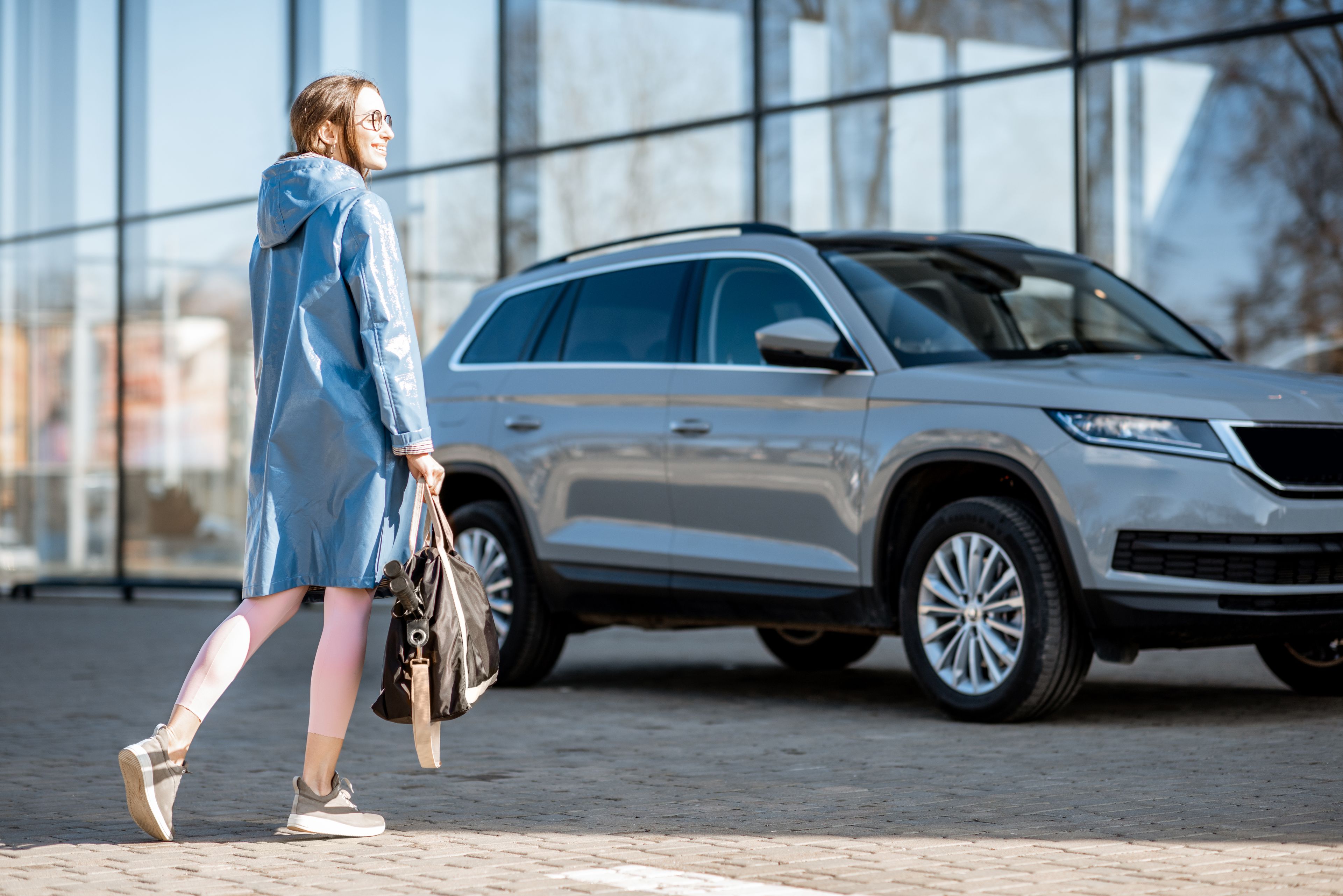 Woman in a light-blue raincoat carrying a black bag walking toward a silver Oscar Car Rental SUV parked by a glass-front building.