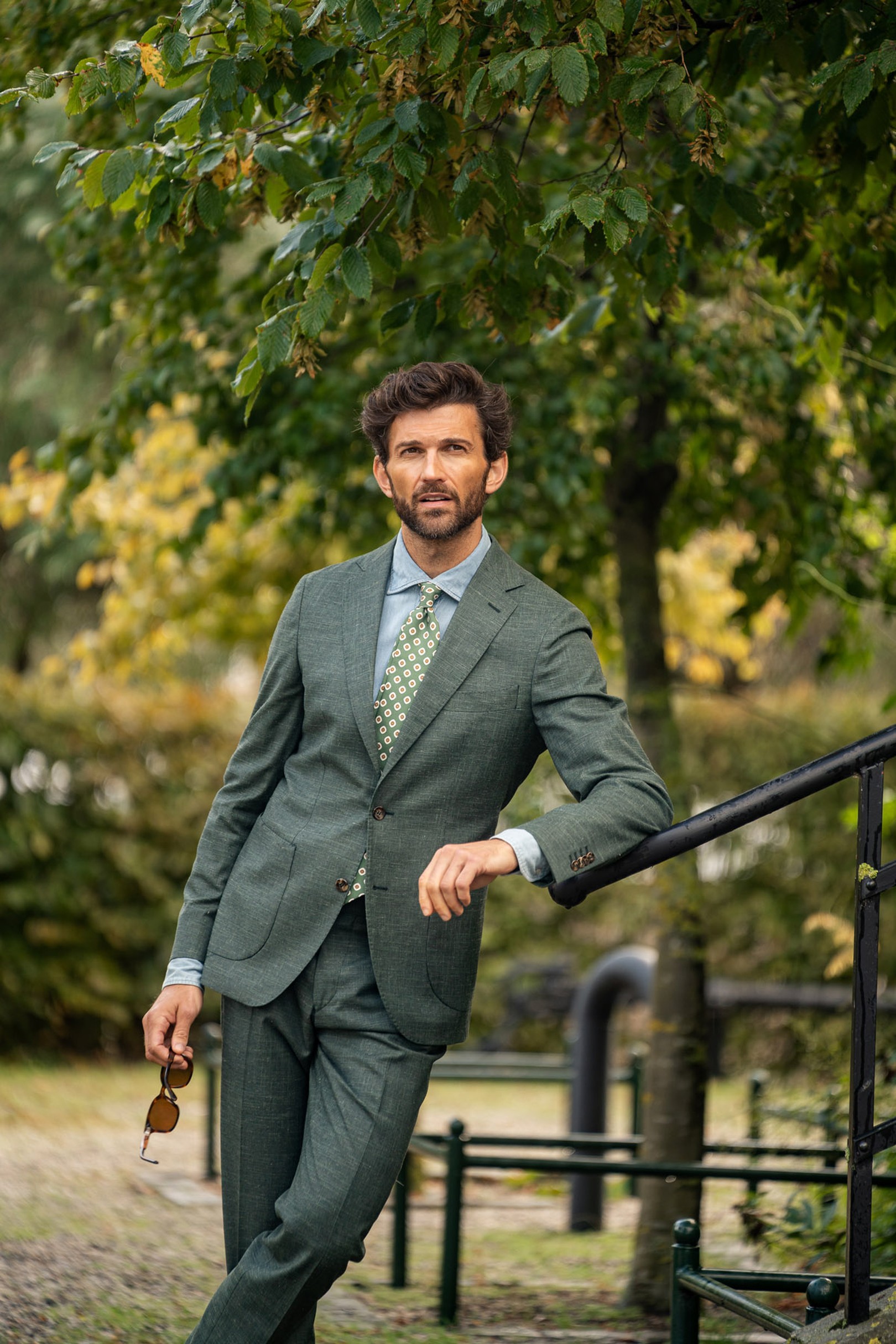 A man in a green suit with a patterned tie stands outdoors near a railing, holding sunglasses, with trees in the background.