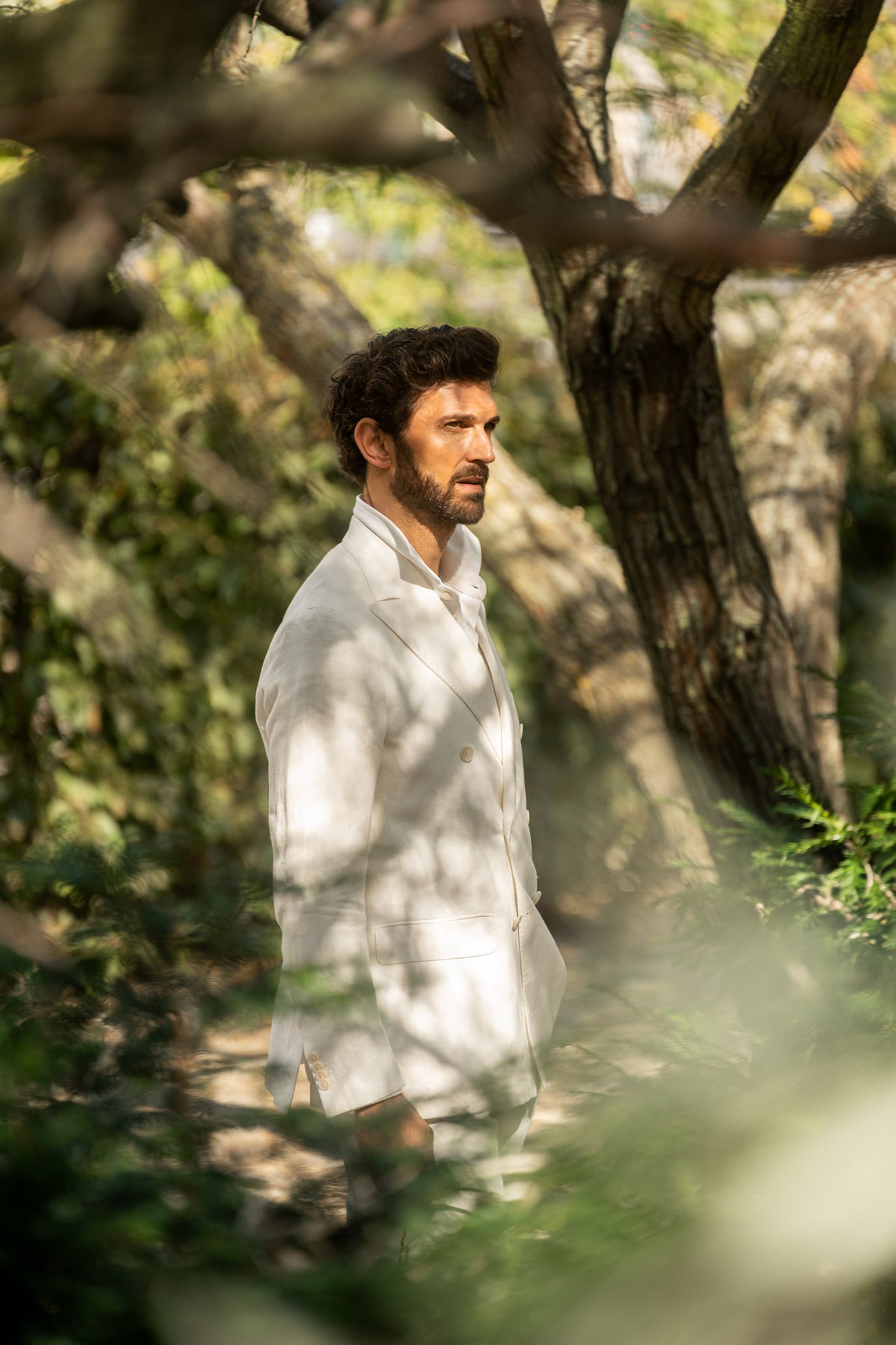 A man in a white suit stands thoughtfully in a lush, sun-dappled forest, surrounded by trees and greenery.