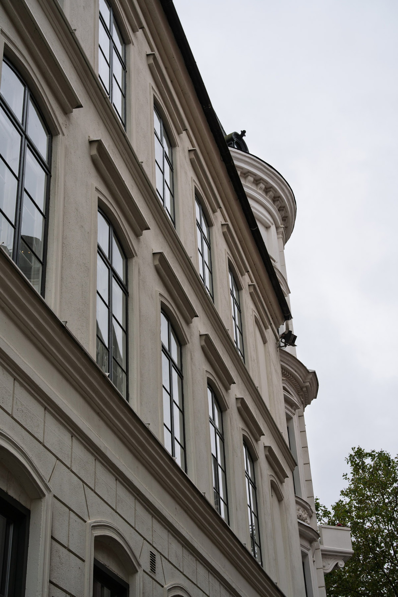 Angled view of a classic European-style building with large arched windows and a cylindrical tower against a cloudy sky.