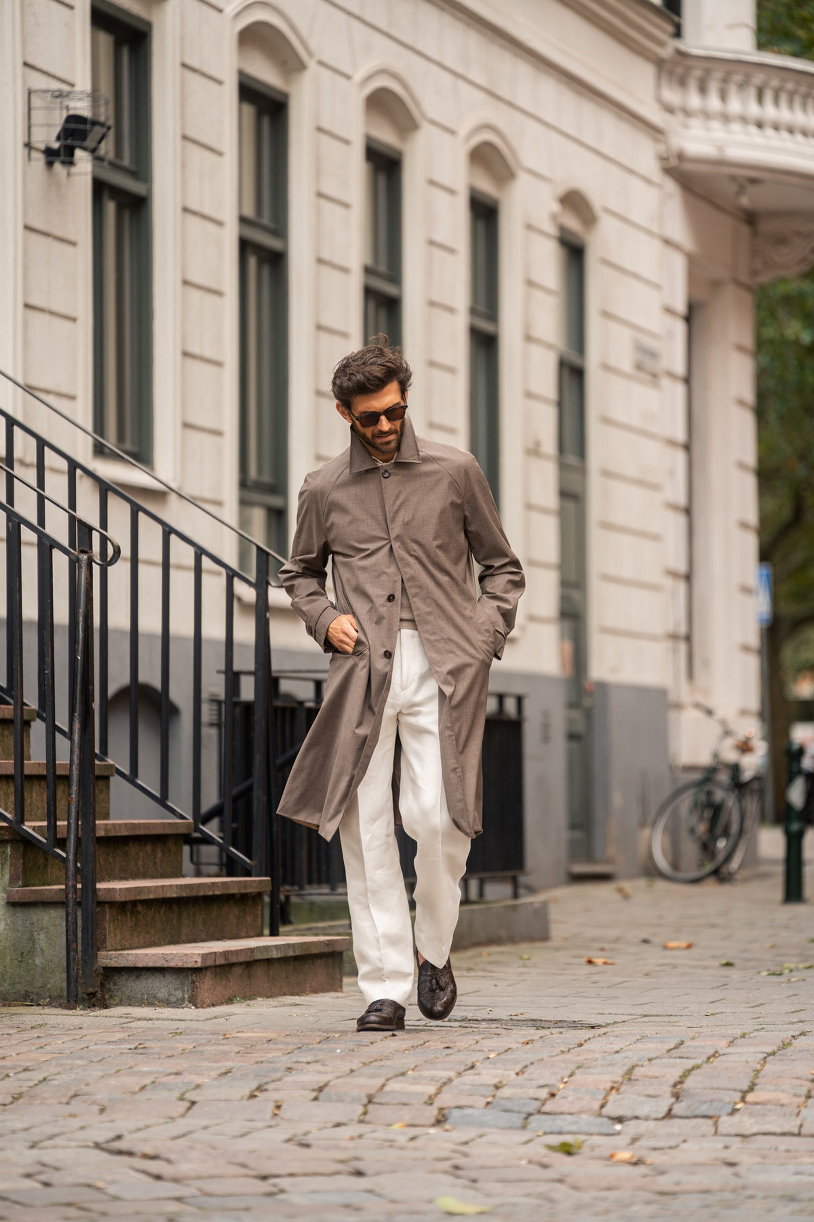 Man in sunglasses and a long coat walks confidently on a cobblestone street, with a bicycle and old building in the background.
