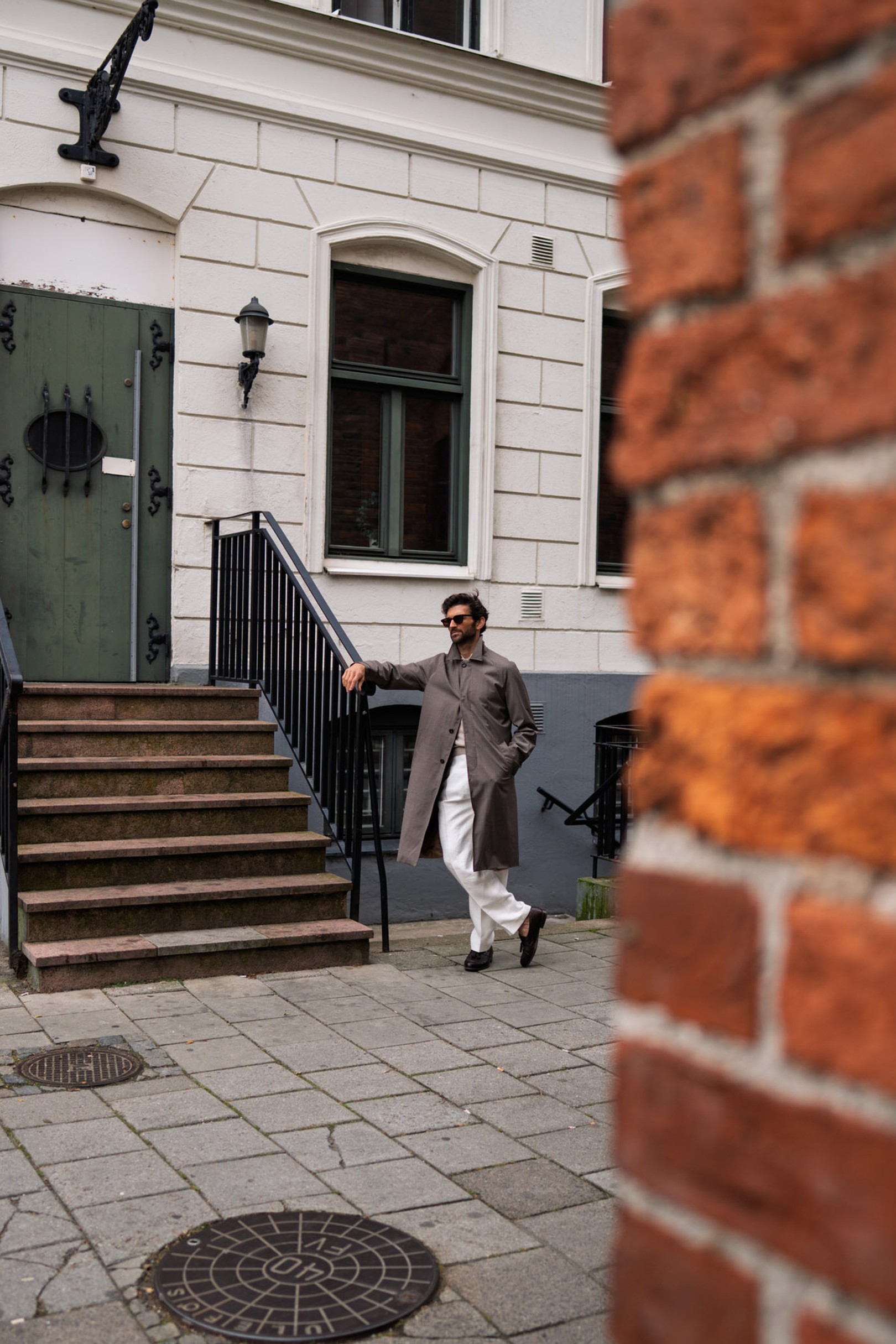 Man in a gray coat and white pants walking down steps outside a building with white facade; partial red brick wall in foreground.