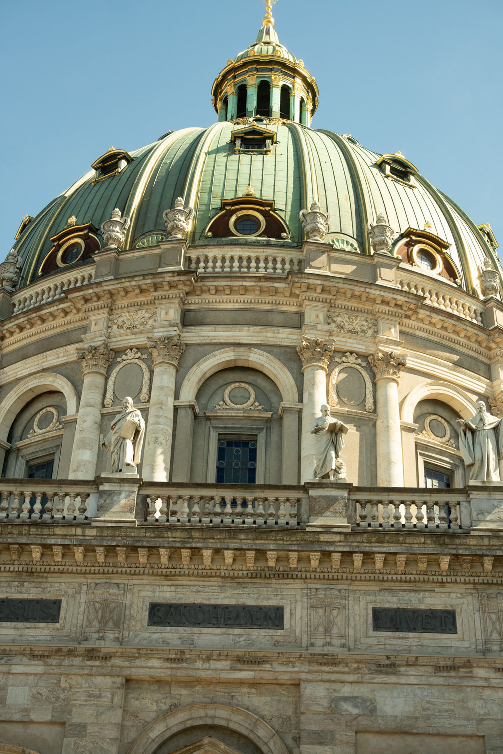 A close-up view of an ornate domed building with statues, decorative columns, and architectural details under a clear blue sky.
