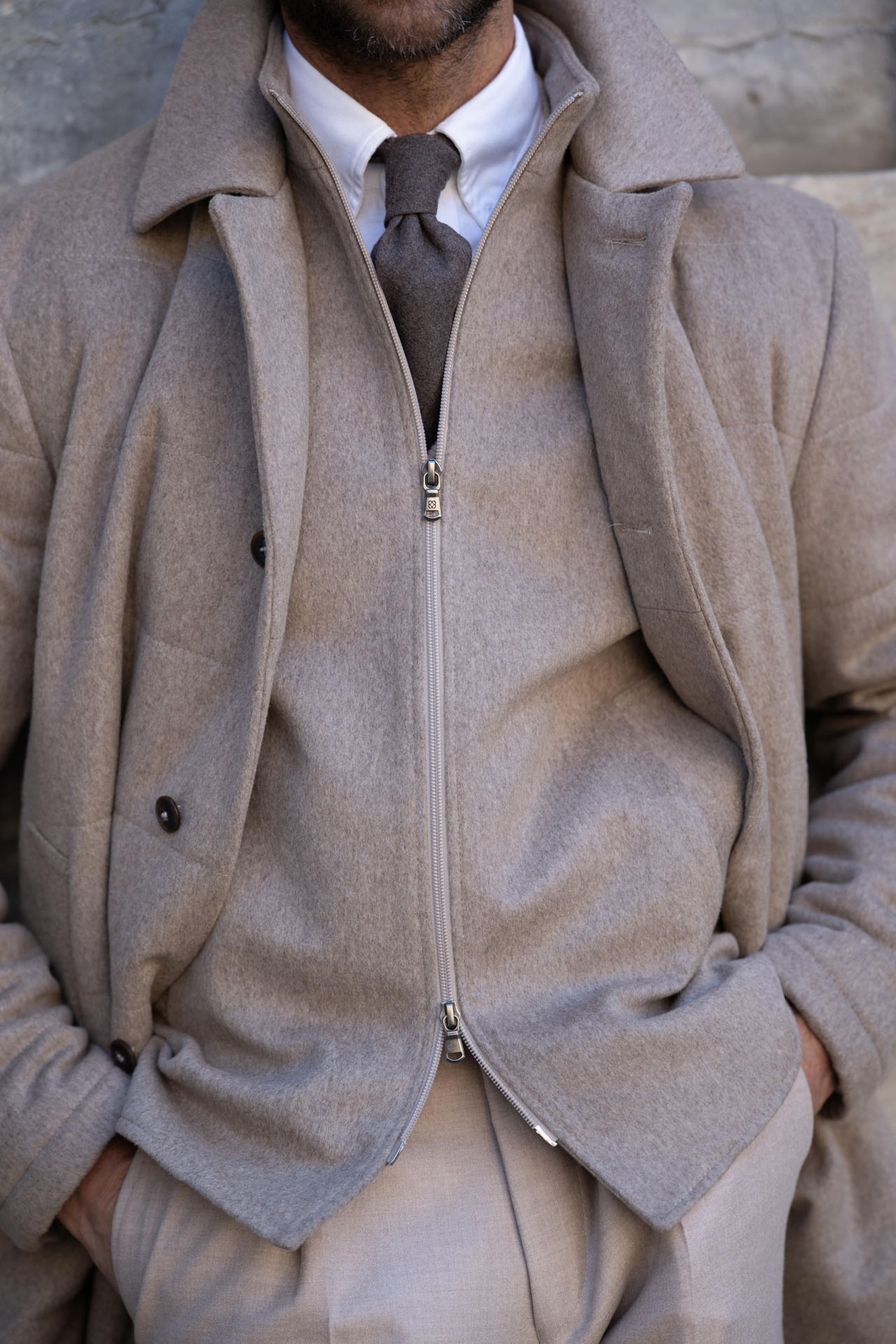 A man in a beige outfit with a zipped jacket, buttoned coat, white shirt, and brown tie, hands in pockets, standing against a stone wall.