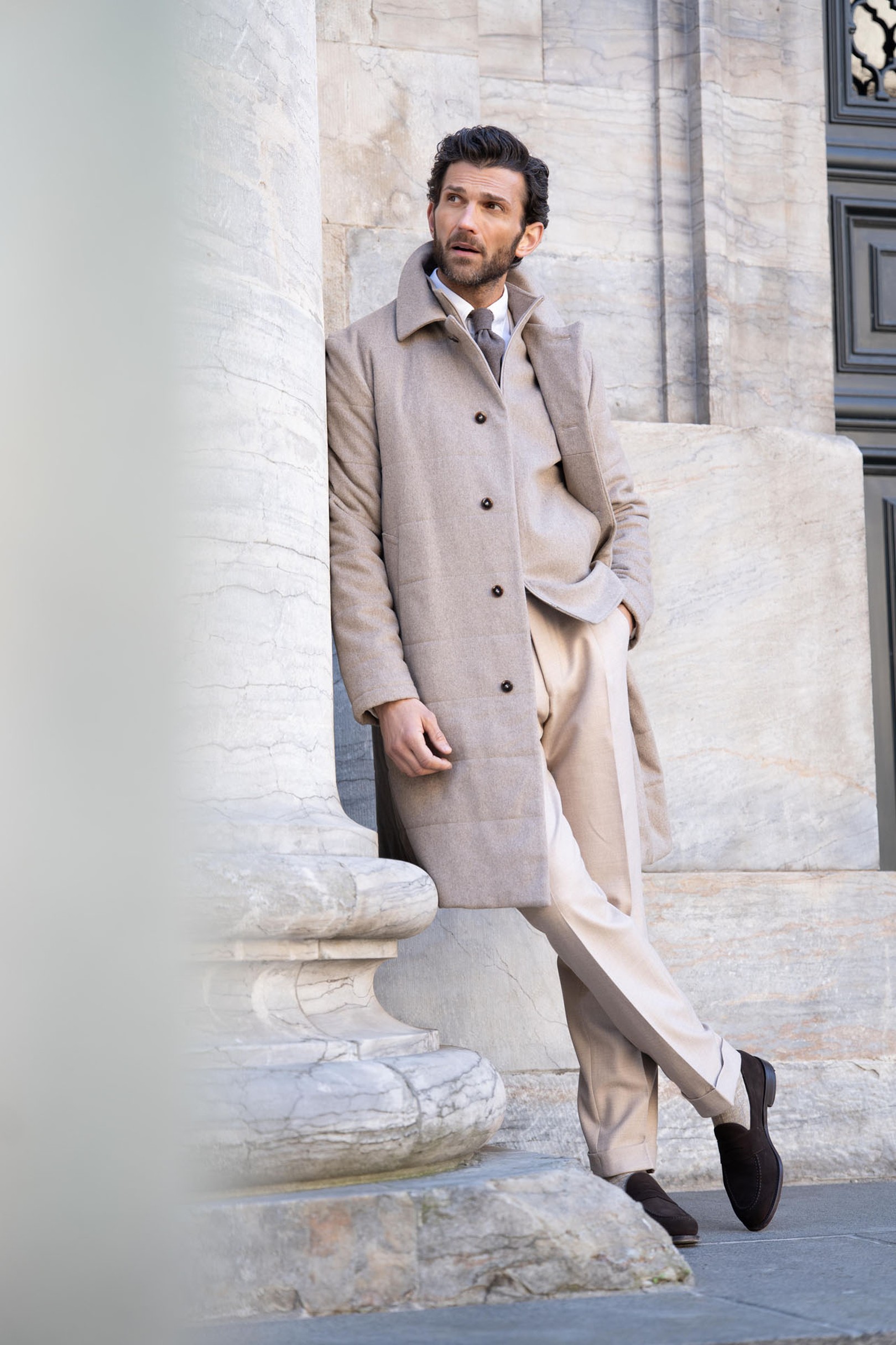 Man in beige coat and trousers leans against a marble column, outdoors, with a thoughtful expression.