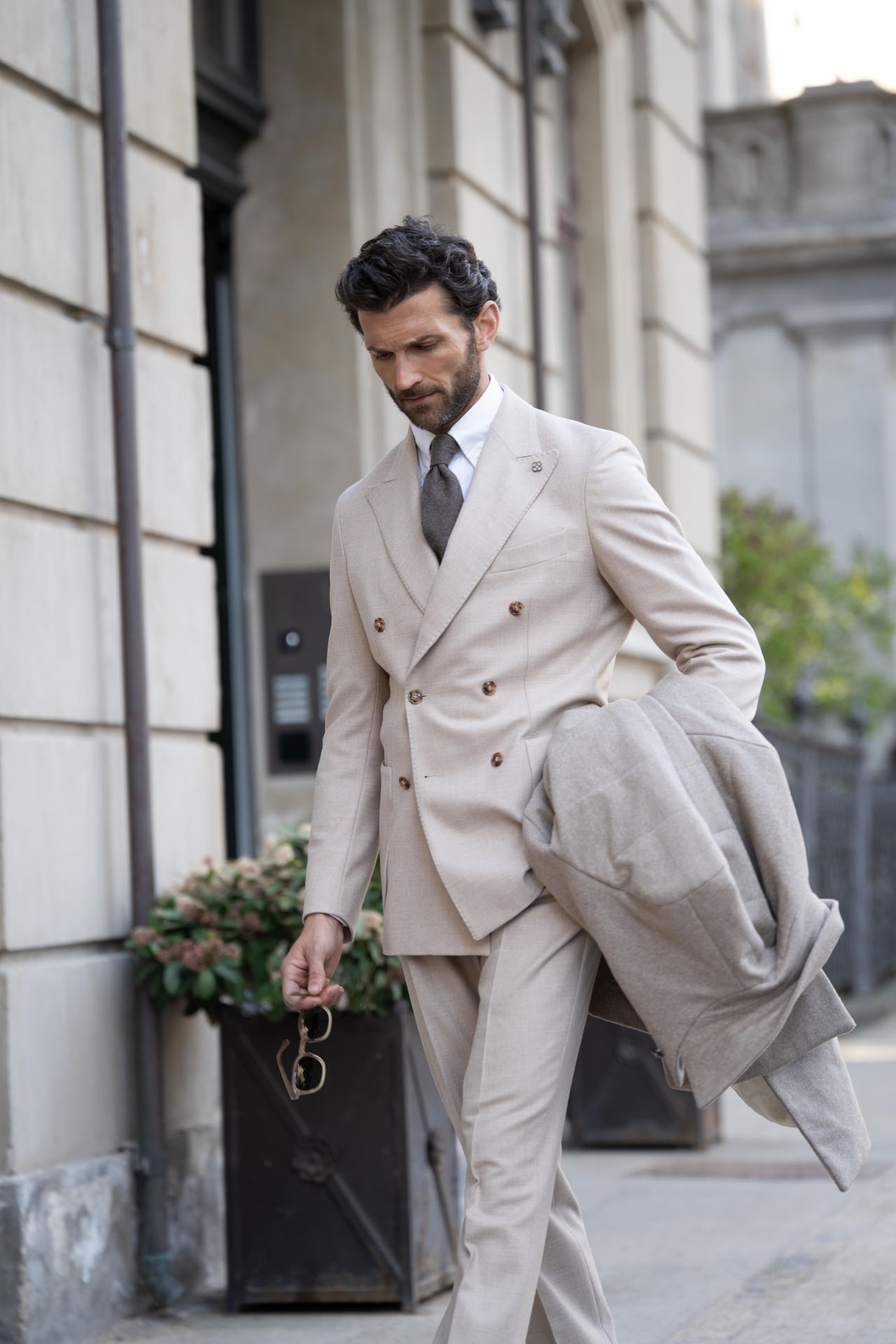 A man in a beige double-breasted suit walks confidently, holding a coat and sunglasses, on a city street lined with elegant buildings.