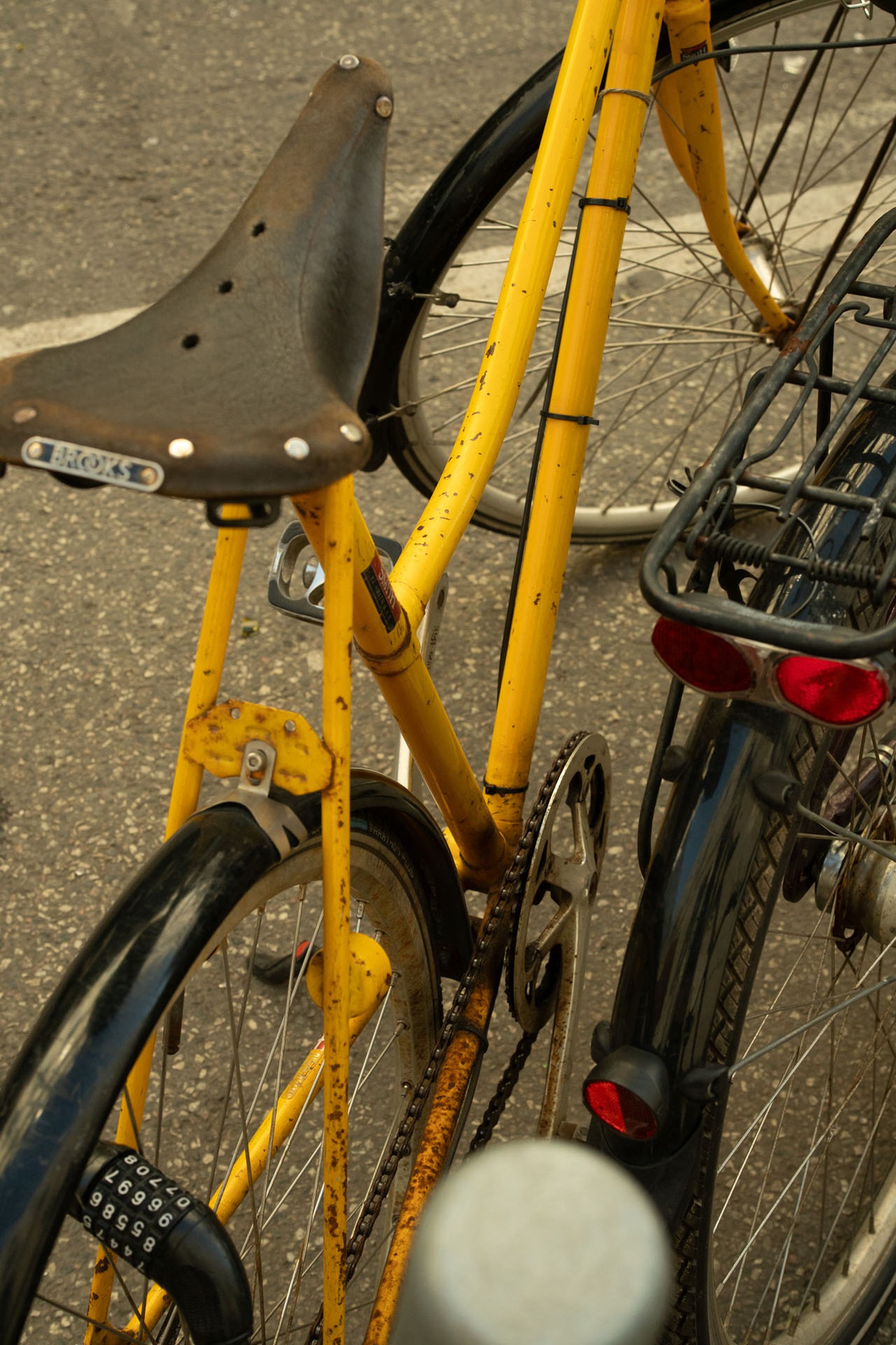 Close-up of a yellow bicycle with a weathered leather saddle and black fenders, parked alongside another bike on an asphalt surface.