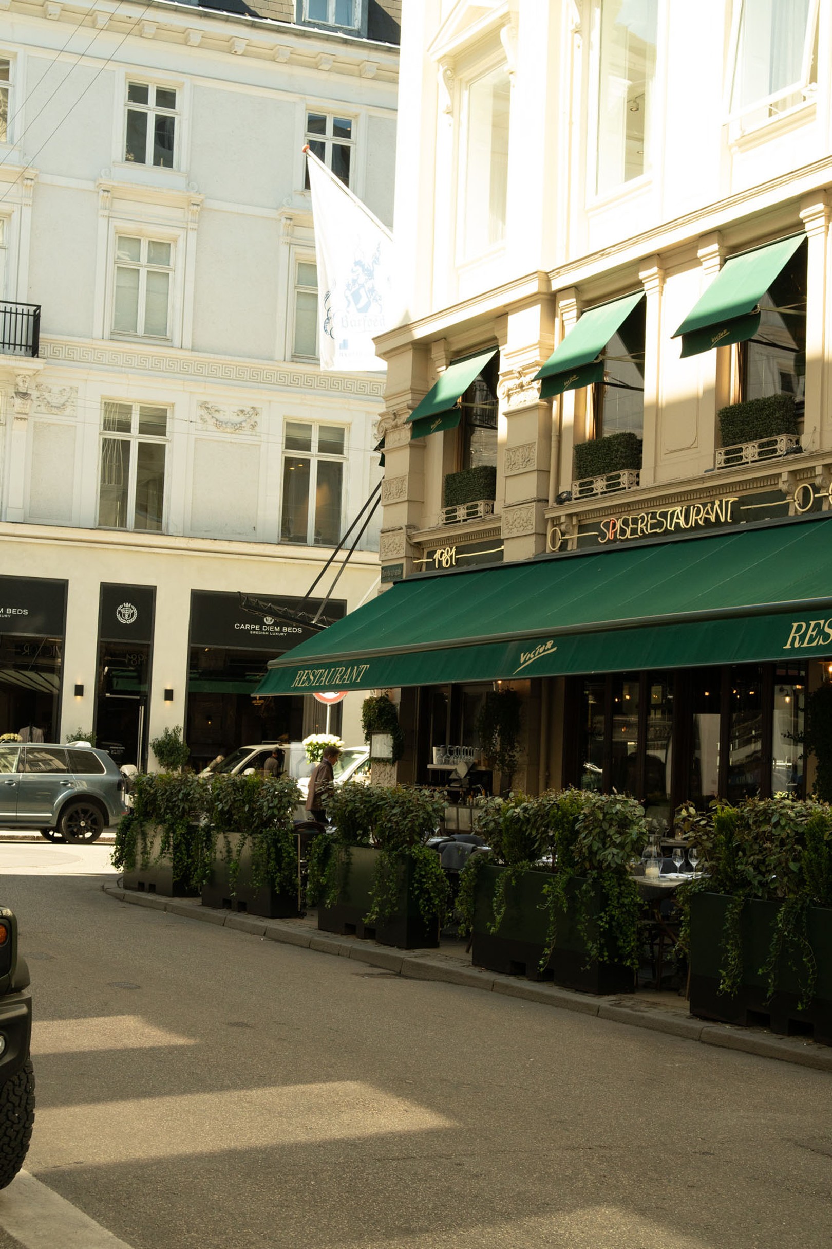 A European street scene with a restaurant featuring green awnings, outdoor seating with plants, and several parked cars nearby.