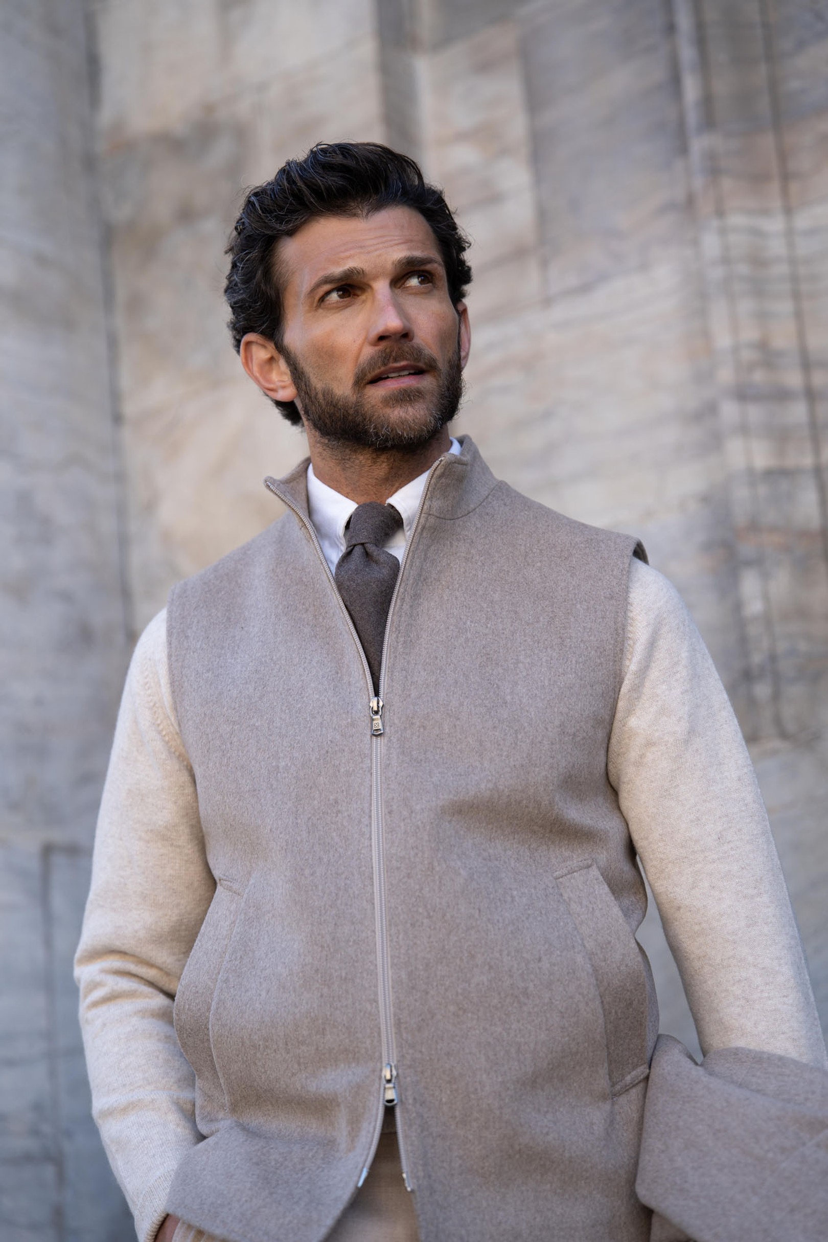 A man with short dark hair and a beard wearing a light gray zip-up vest over a cream shirt and tie, standing against a stone wall.