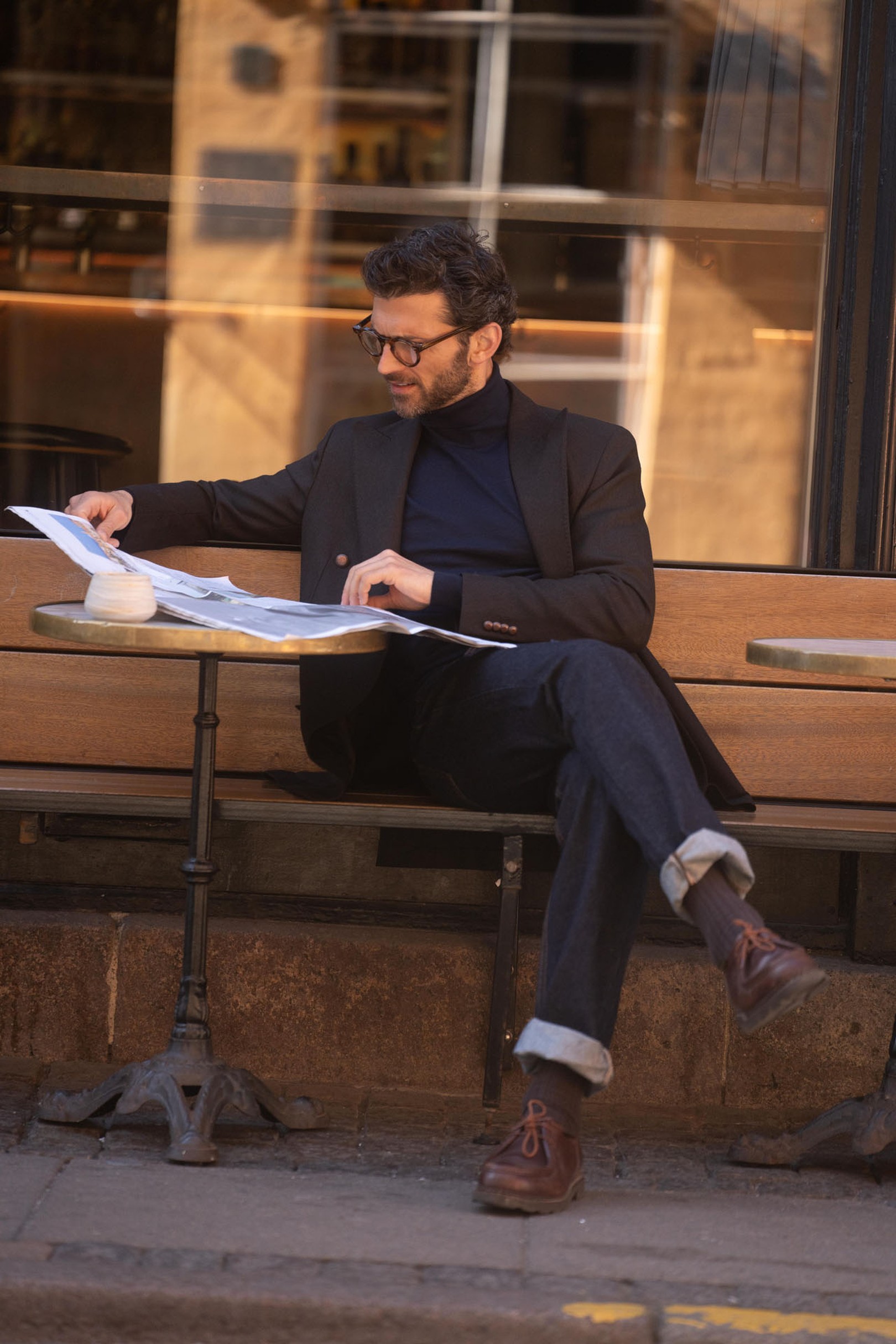 Man sitting at an outdoor café table, reading a newspaper. He wears glasses, a dark blazer, and rolled-up jeans, with brown shoes.