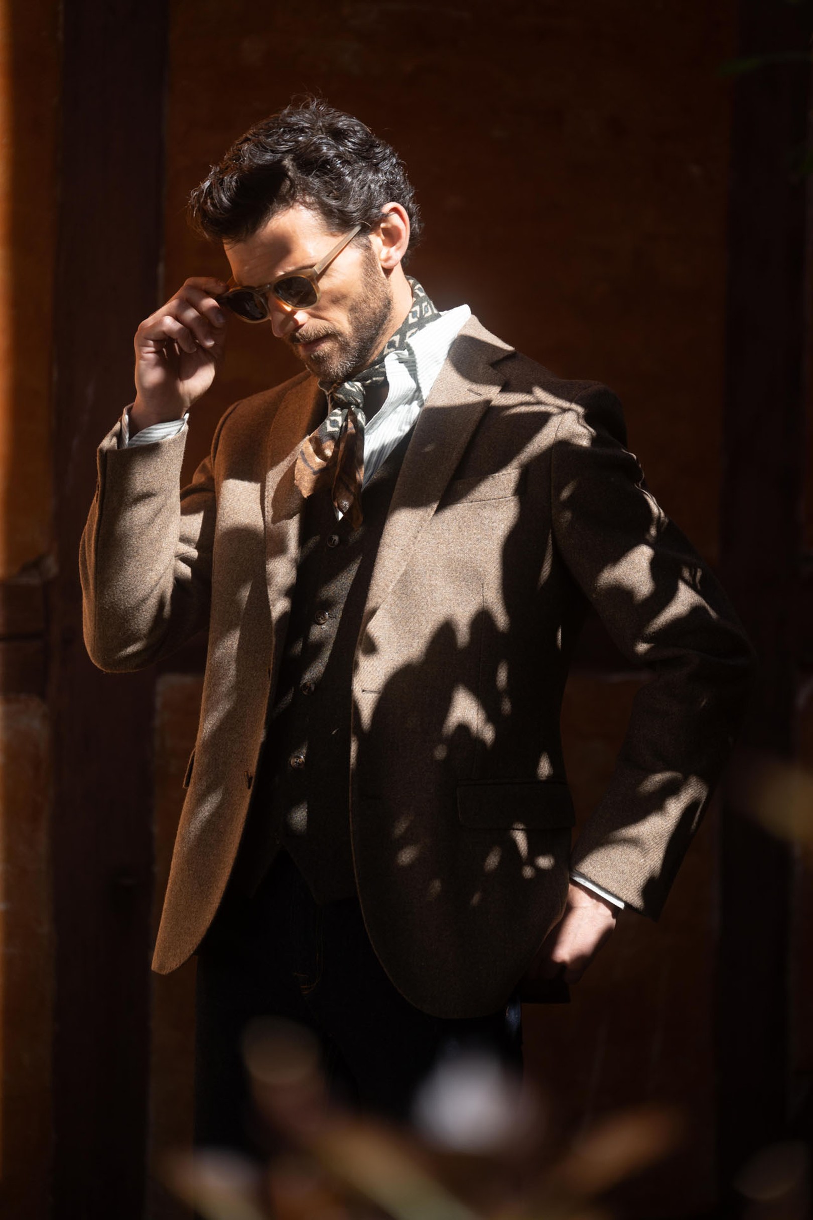 A man in a brown suit and sunglasses stands in partial shadow, adjusting his glasses and wearing a patterned scarf inside.