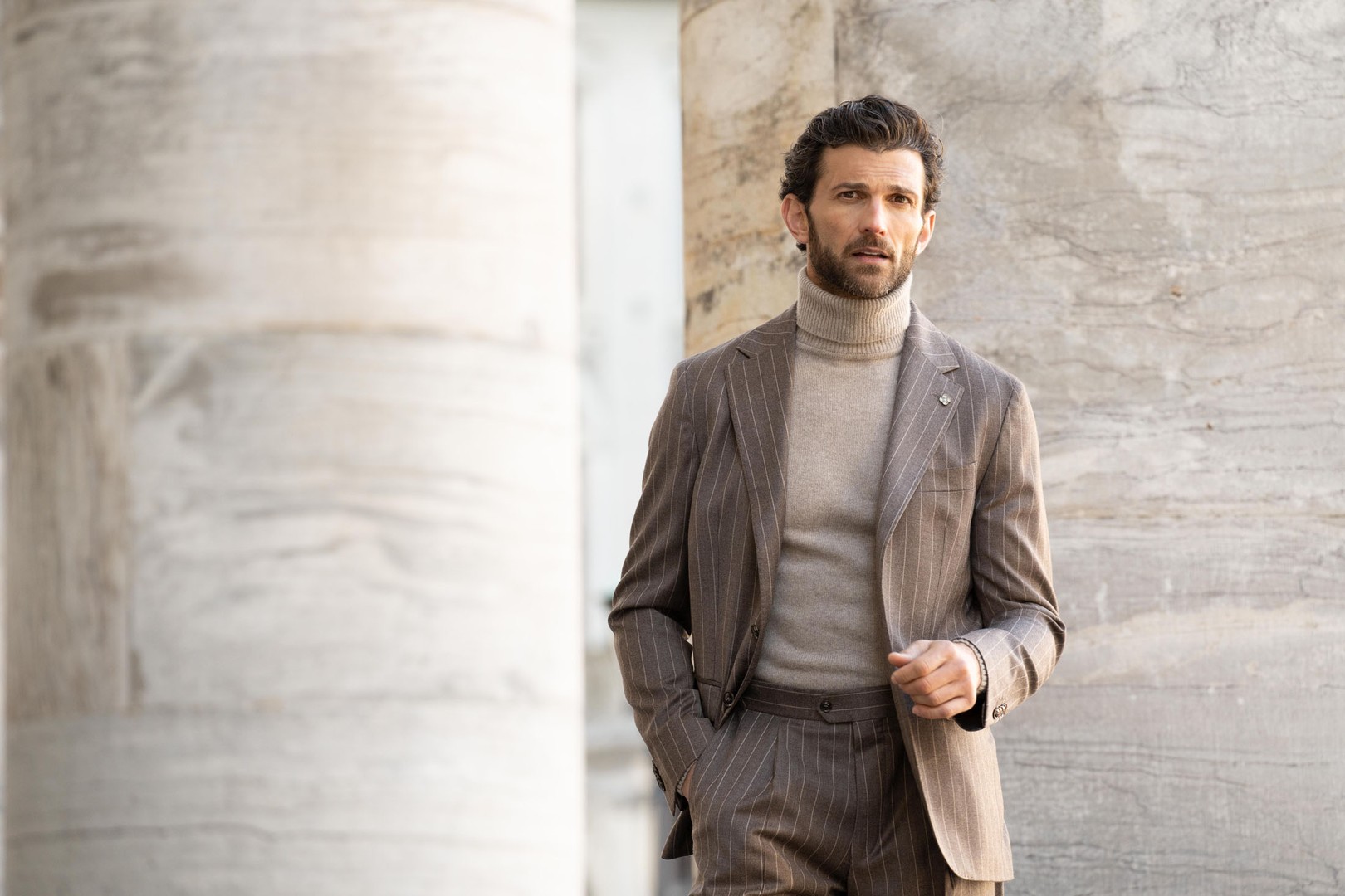A man in a brown pinstripe suit with a beige turtleneck stands against a marble wall, looking thoughtfully to the side.