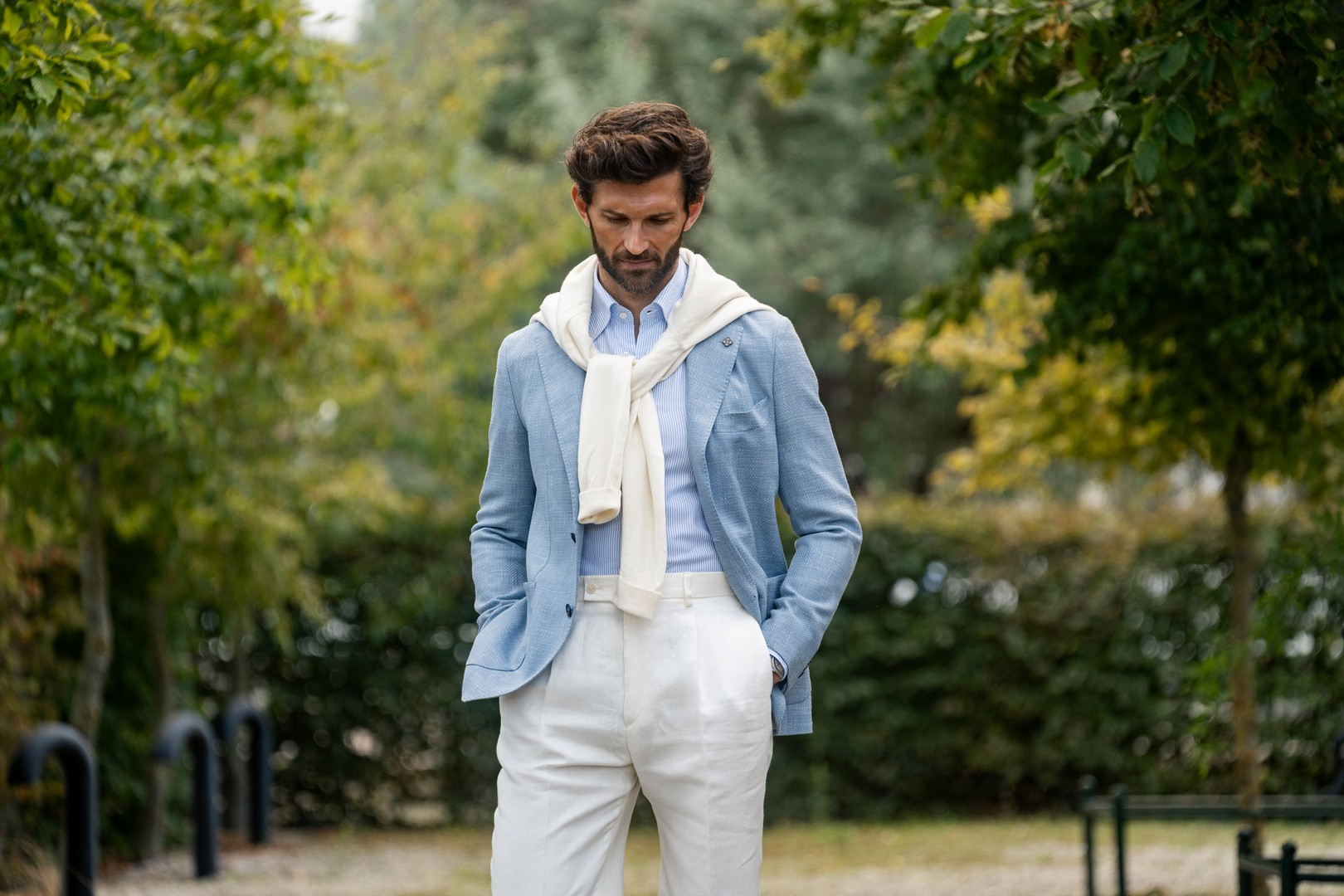 Man in a light blue blazer with a white sweater draped over his shoulders, walking outdoors with hands in pockets, surrounded by greenery.