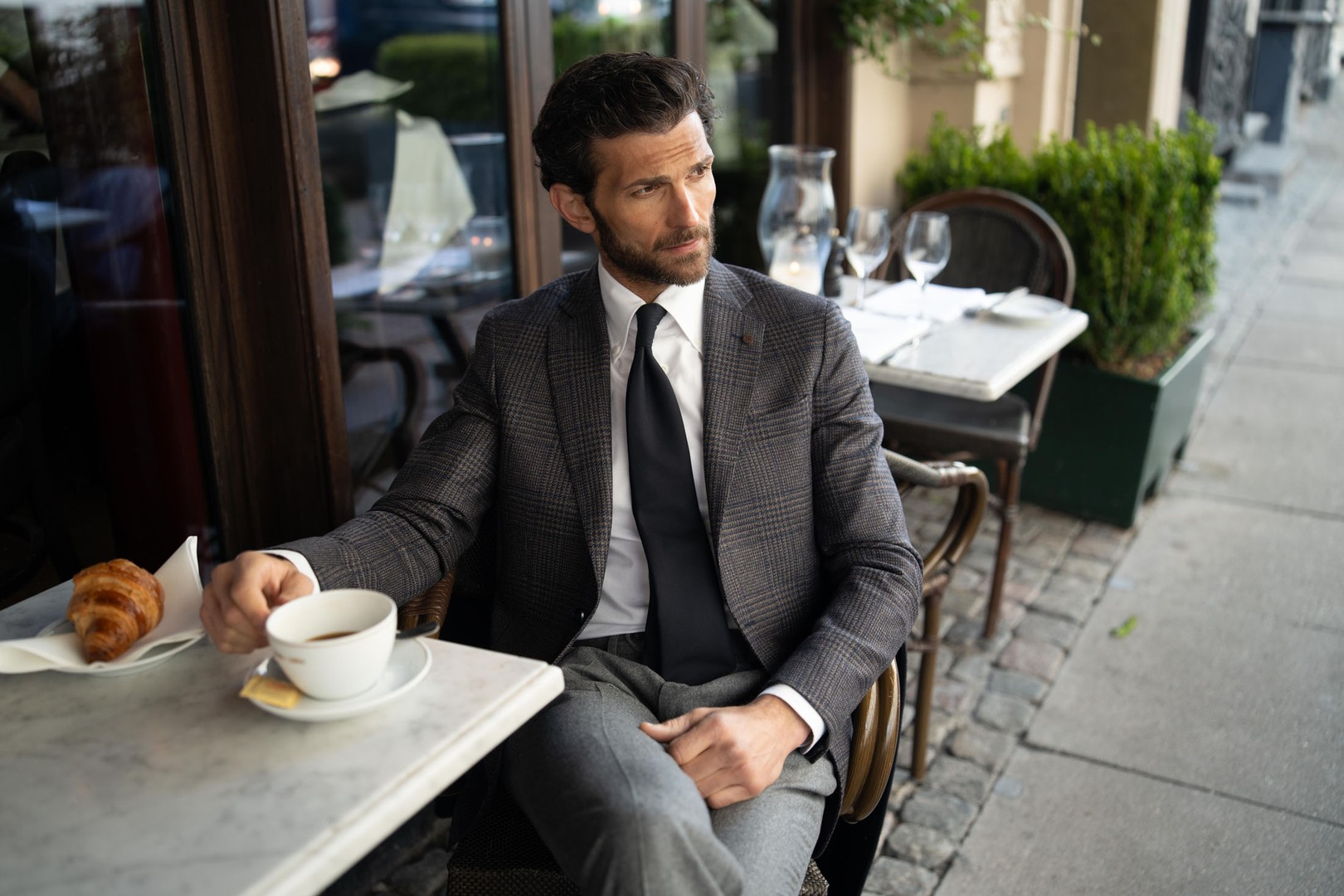 A man in a suit sits at an outdoor cafe table, holding a cup of coffee with a croissant on the table, looking contemplative.