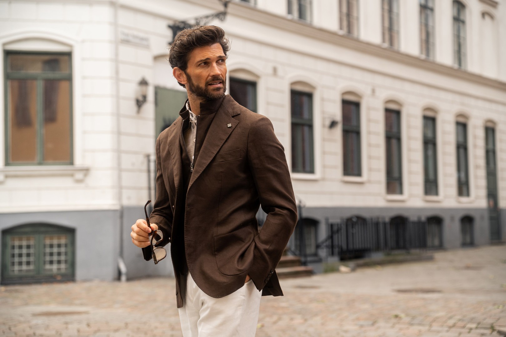 A man in a brown blazer and white pants stands on a cobblestone street holding sunglasses, with buildings in the background.