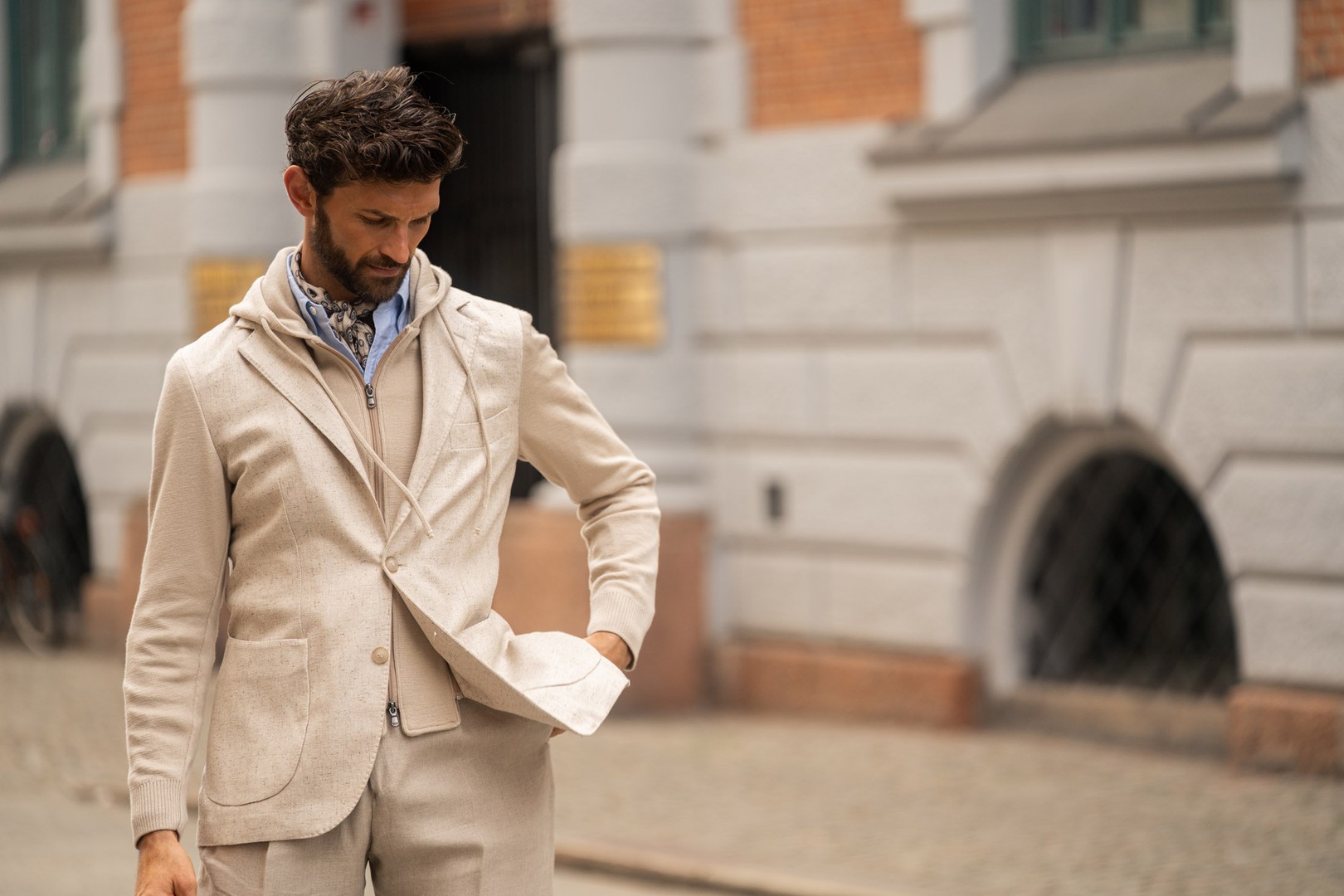 Man in a beige suit and hoodie stands outdoors against a brick and stone building, looking down with one hand in his pocket.