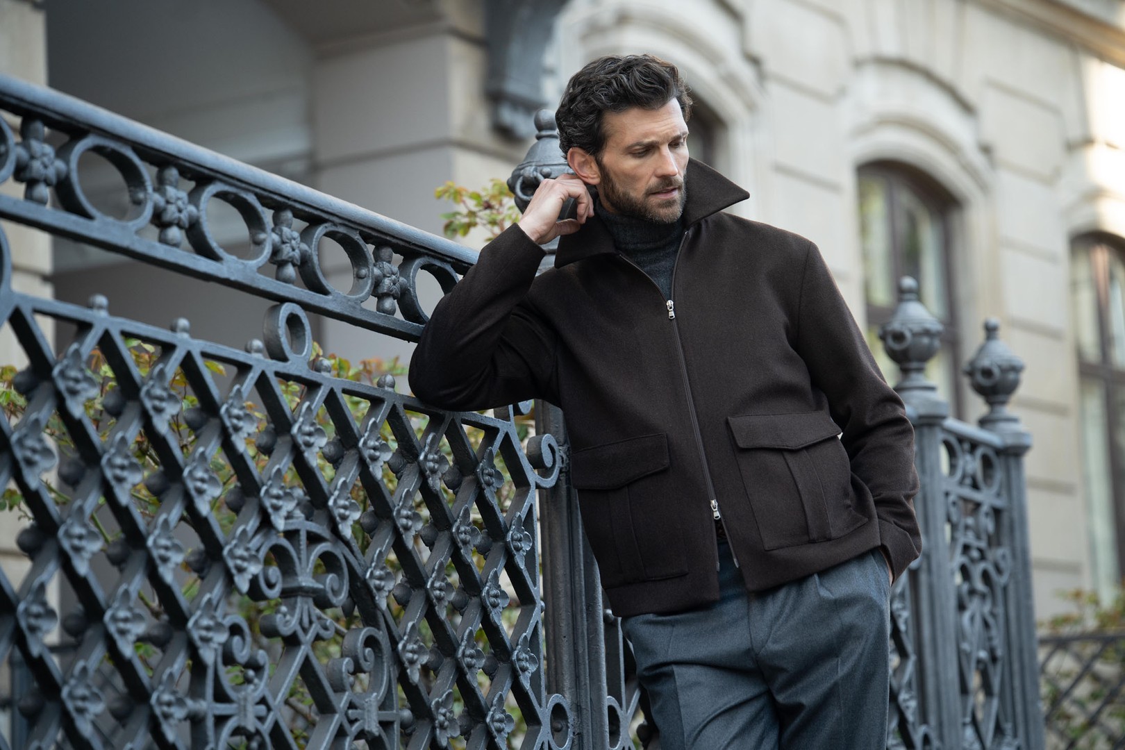 Man leaning against an ornate metal fence, wearing a dark jacket and trousers, looking thoughtfully to the side.