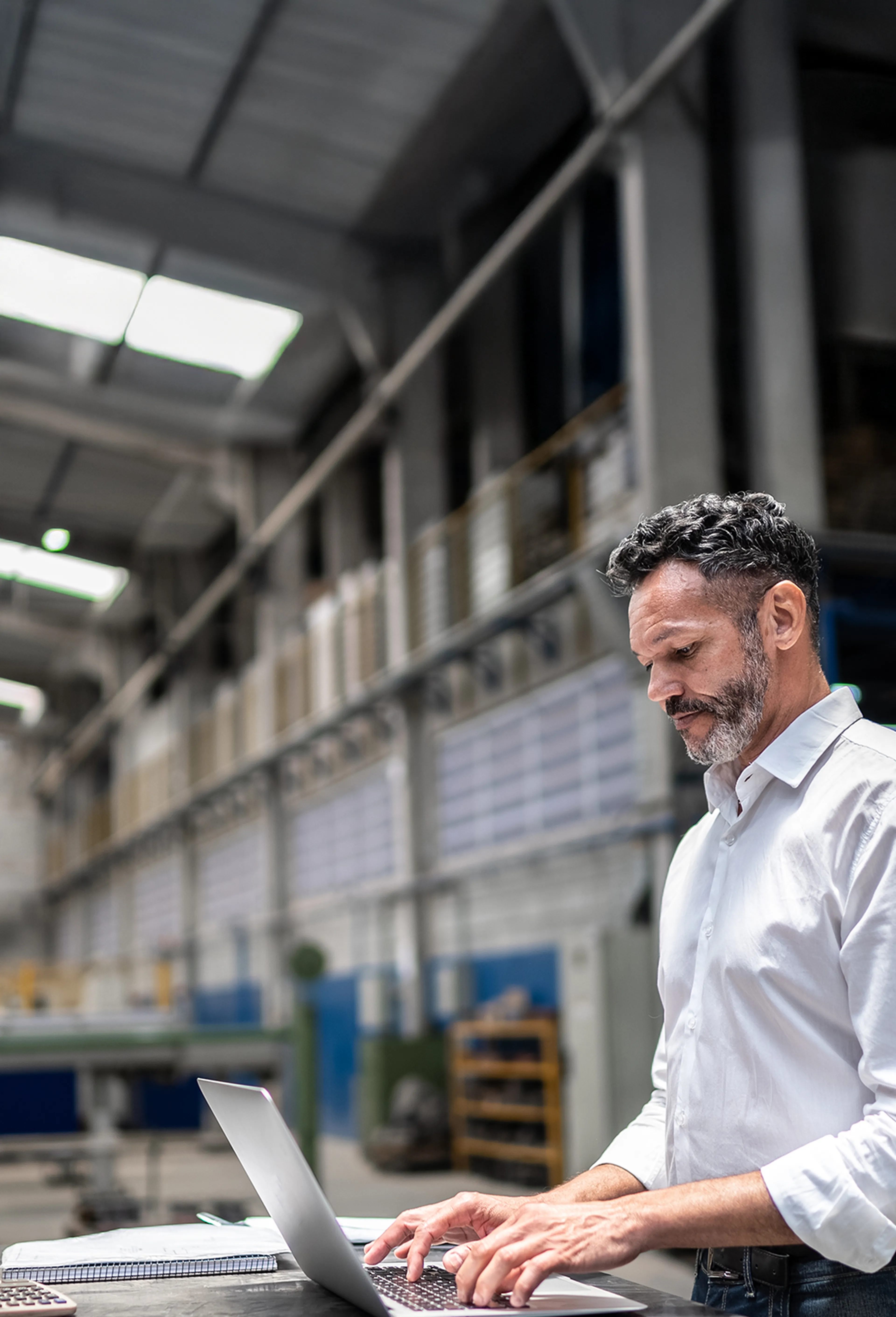 Man in a white shirt working on a laptop in a spacious industrial warehouse with high ceilings and metal structures.