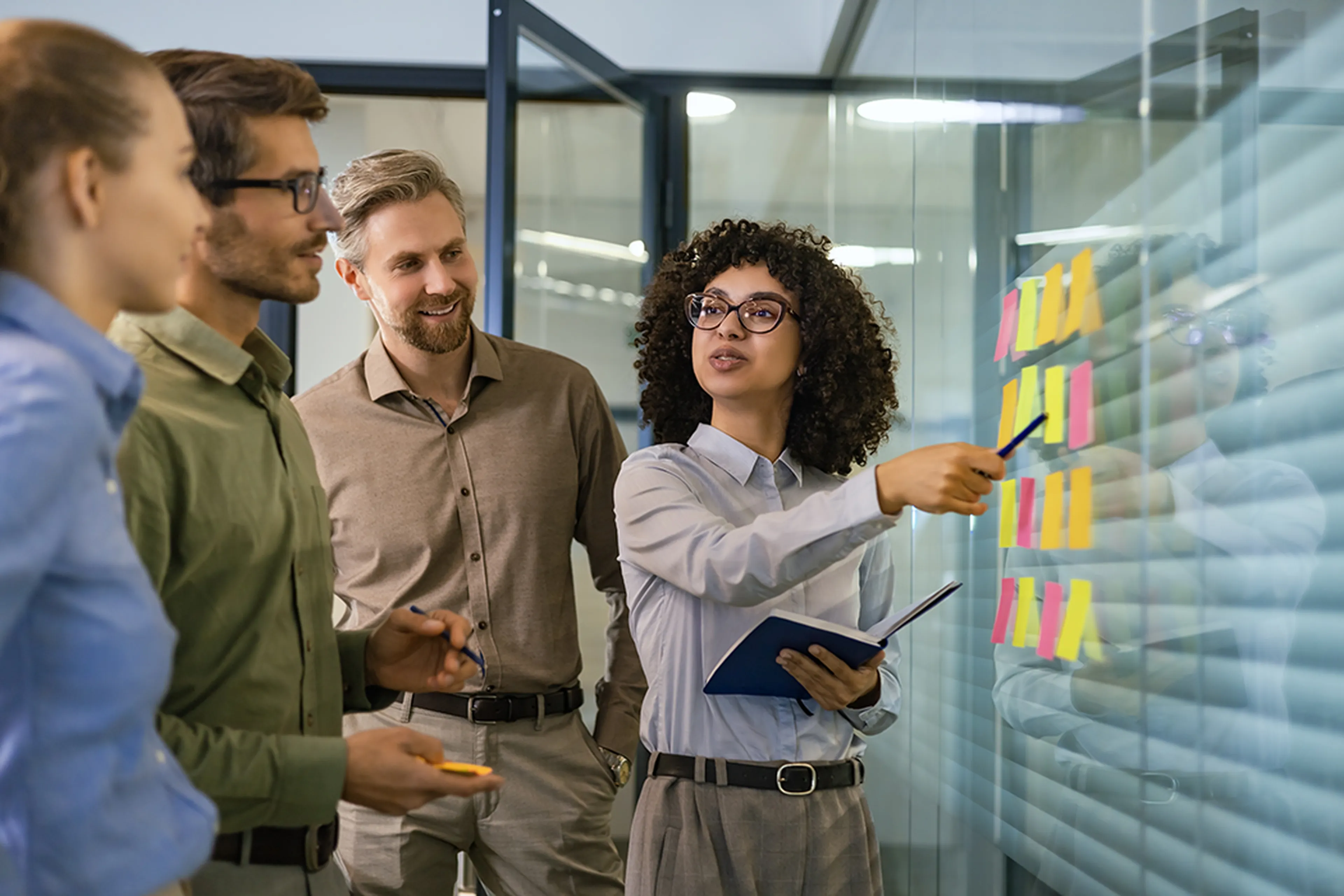 Four colleagues discussing ideas, with a woman pointing at sticky notes on a glass wall. They appear engaged and collaborative.