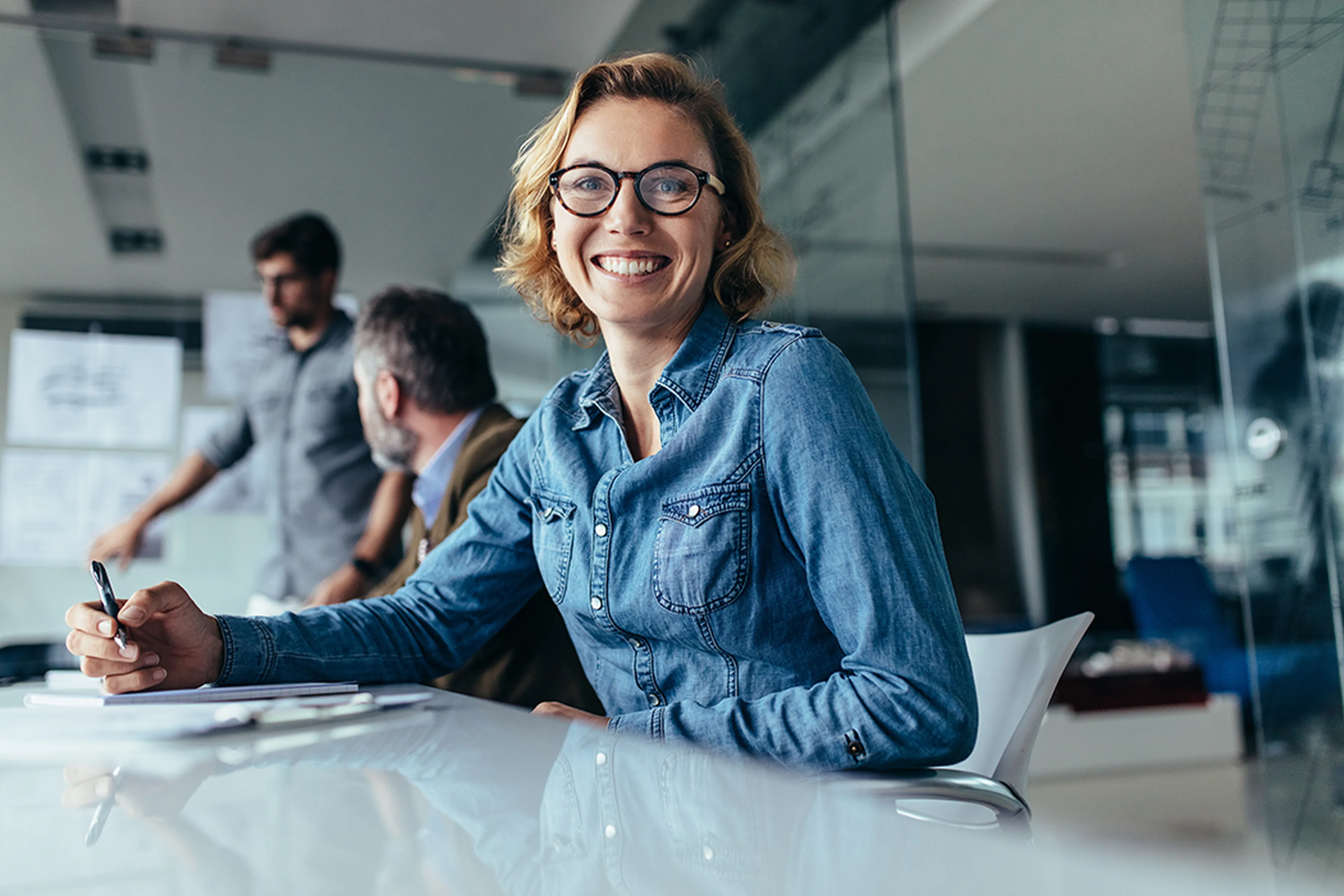 Smiling woman in glasses and denim shirt sits at a conference table, holding a pen. Two colleagues are blurred in the background.