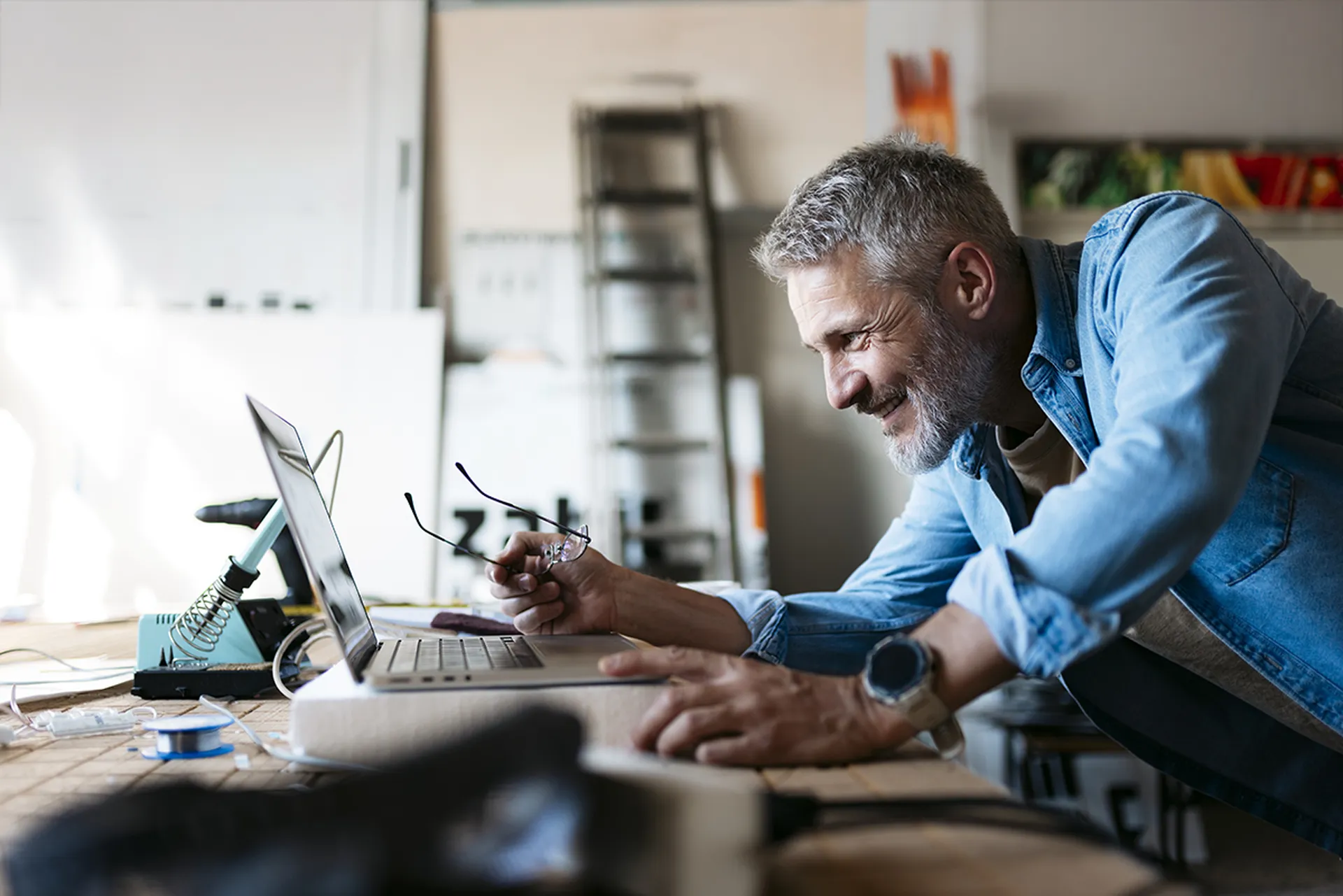 Man smiles while working on a laptop at a cluttered desk in a bright room.