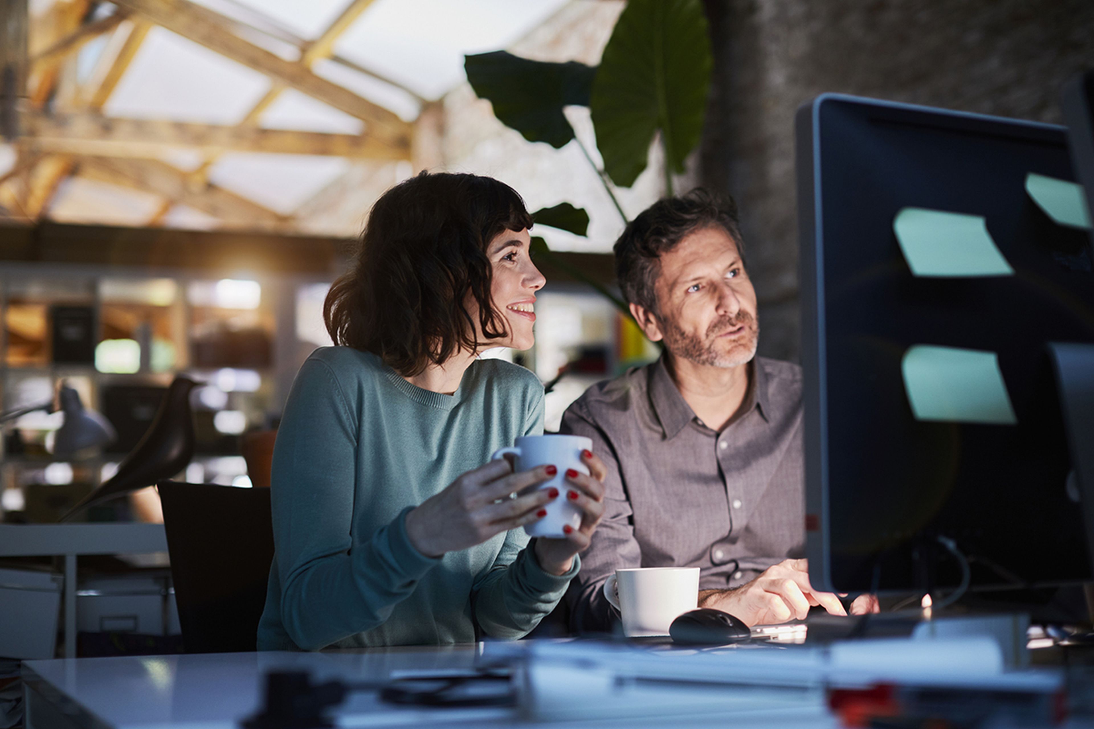 Two people work at a computer in a modern office, with one holding a mug. Natural light filters through large windows, creating a cozy atmosphere.