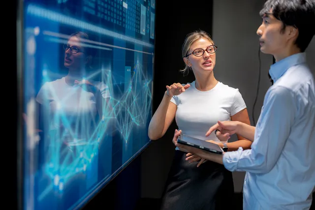 Two people discussing data on a digital screen displaying graphs and charts. One holds a tablet.