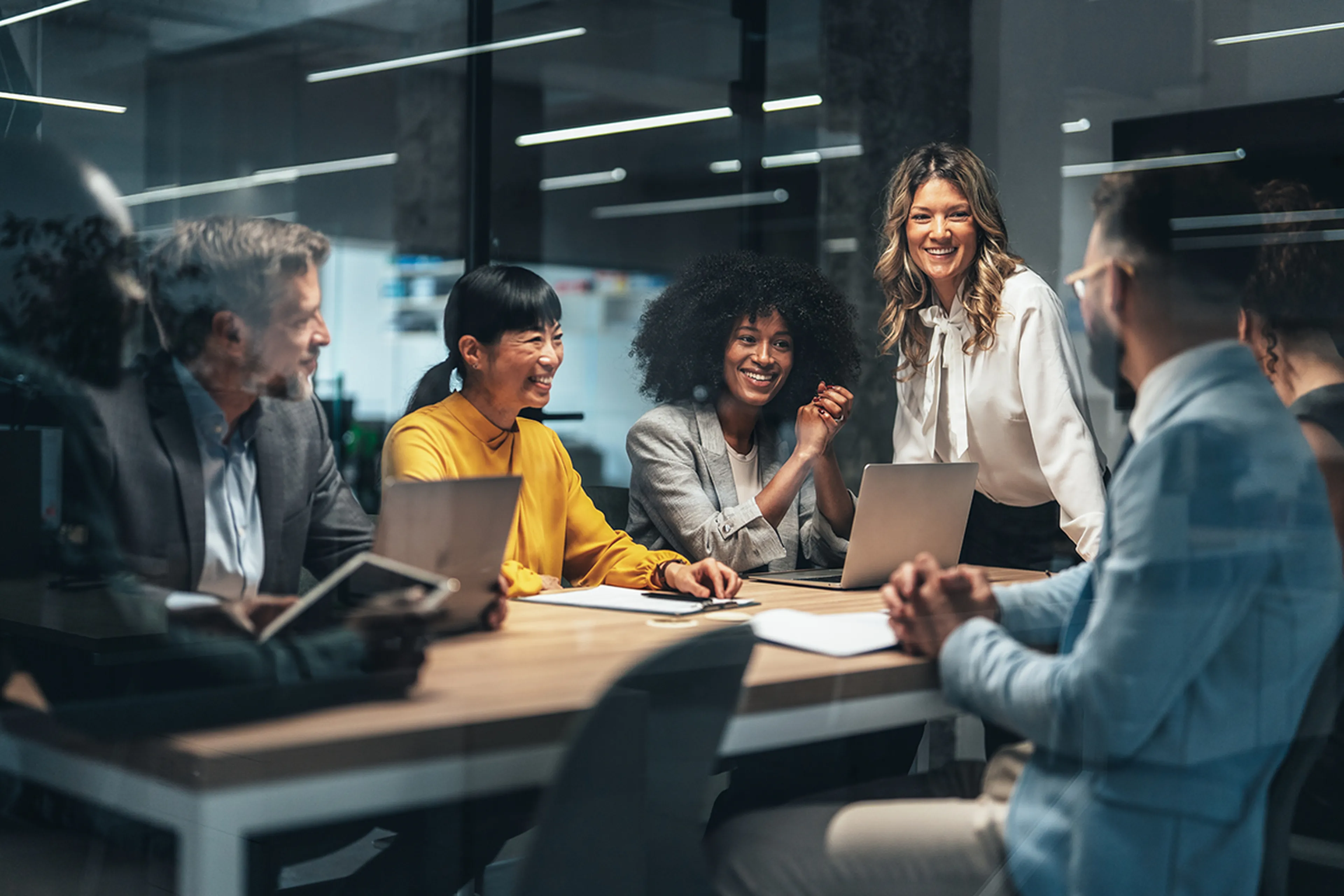 A diverse group of six people in business attire are smiling and discussing around a table in a modern office setting.