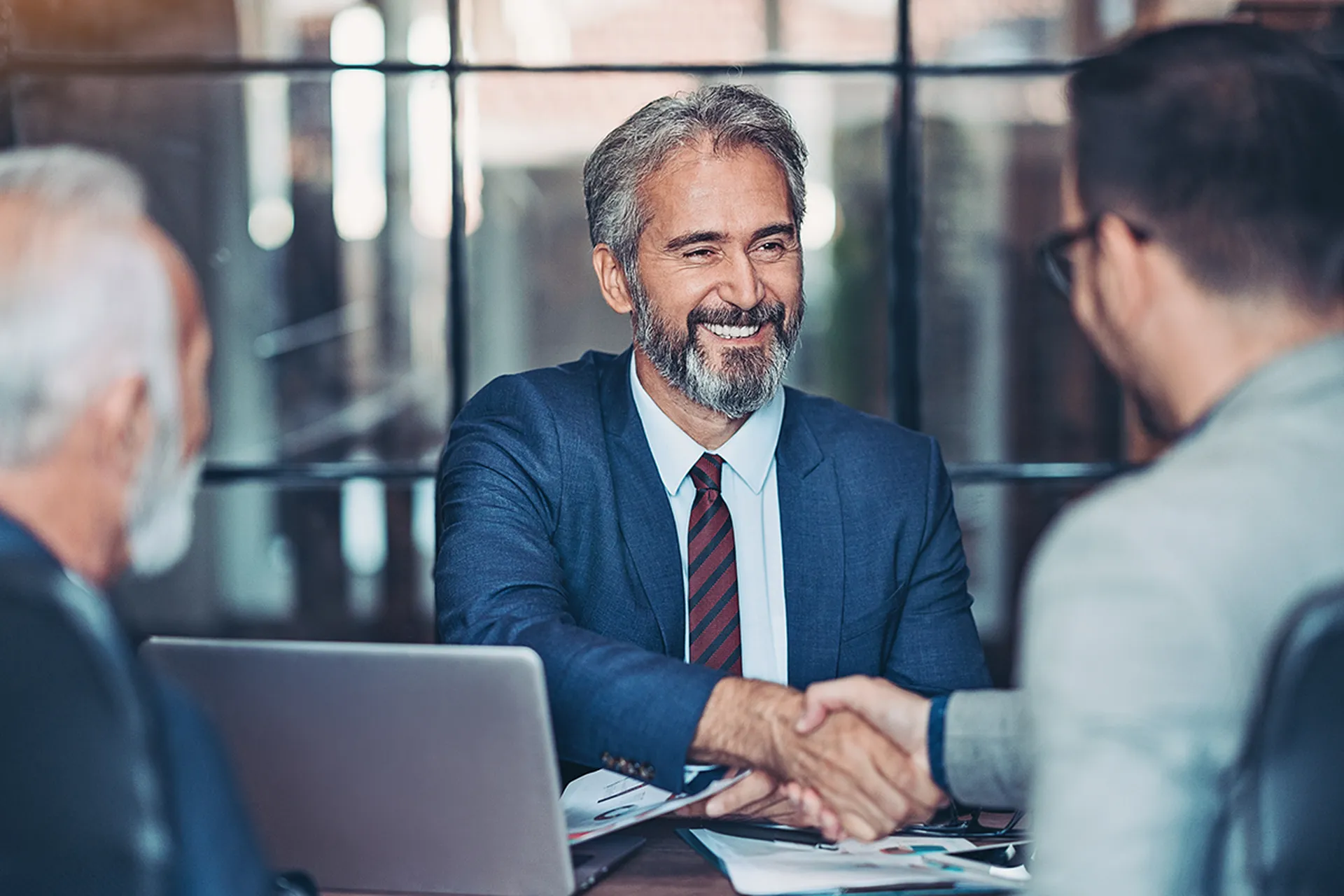 Smiling businessman in a suit shakes hands with a colleague at a meeting table, with a laptop and documents in front of them.