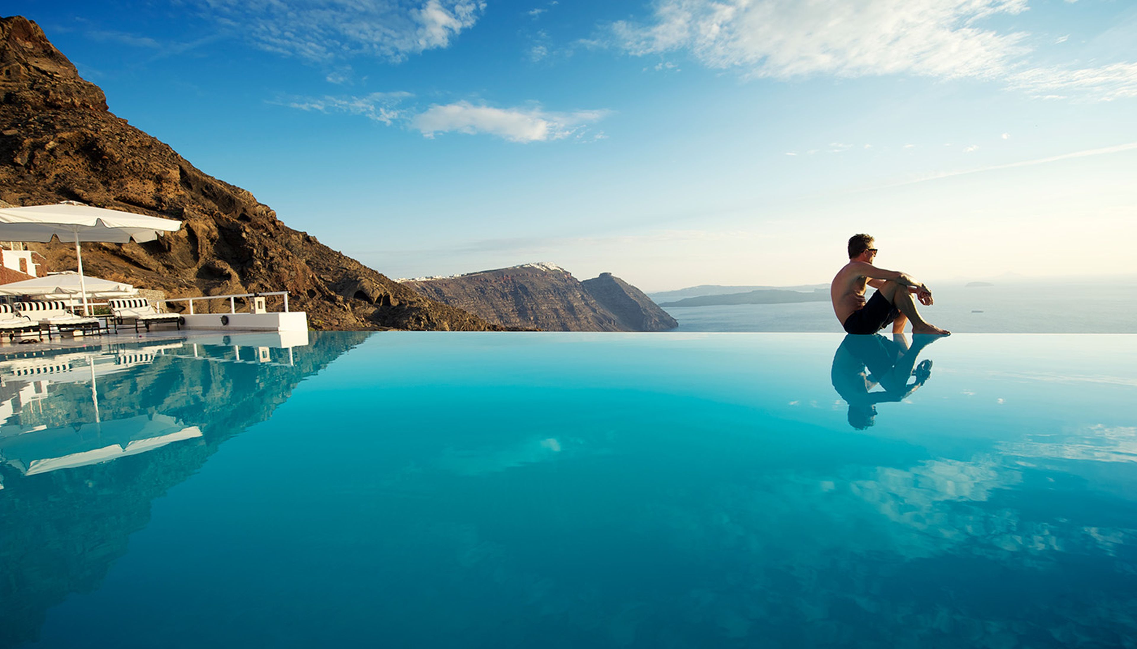 Man seated beside a luxury resort infinity pool with panoramic views of the Aegean sea and coastline.