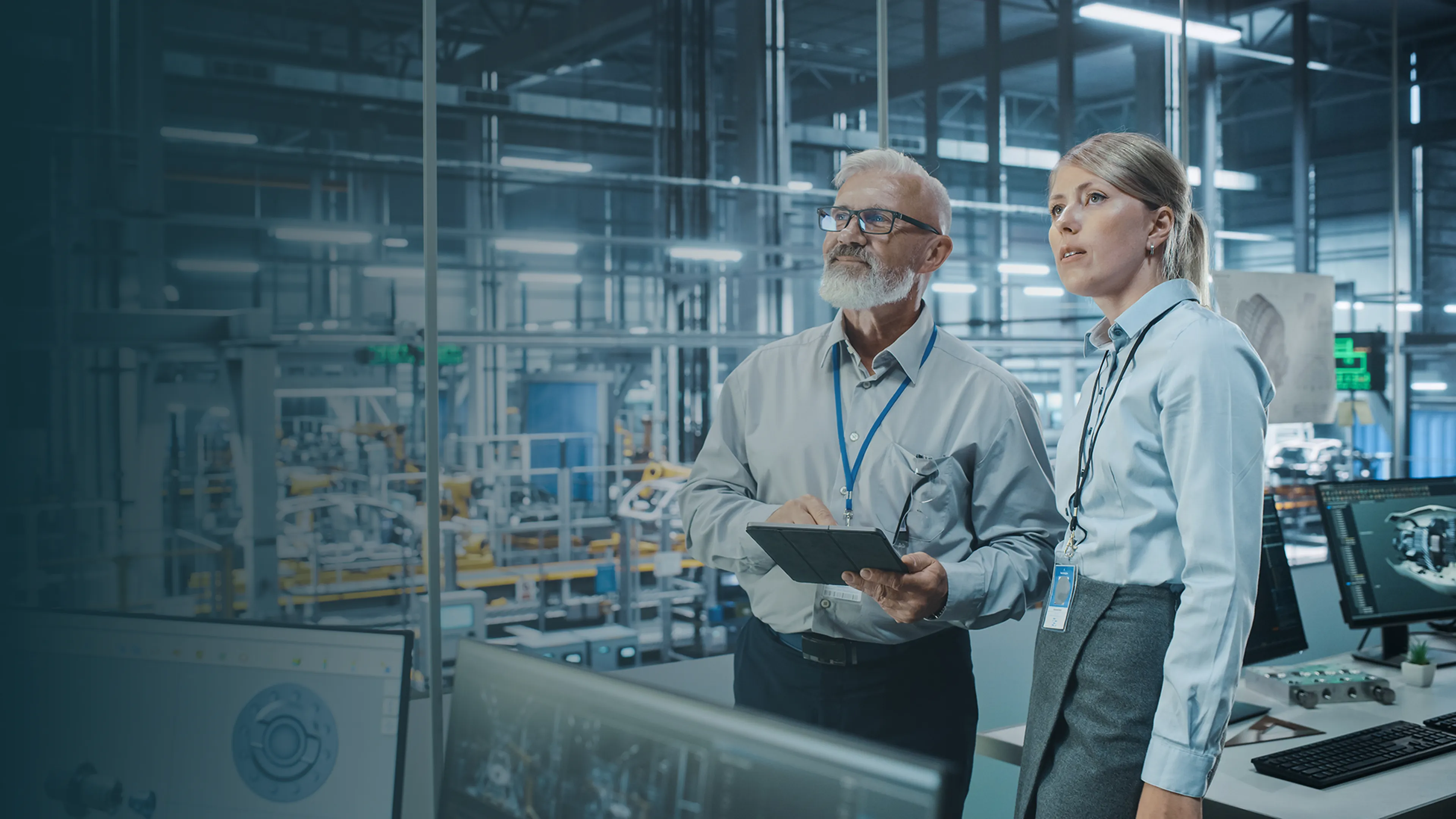 Two professionals in a modern industrial facility discuss while looking at a tablet, surrounded by machinery and computer screens.