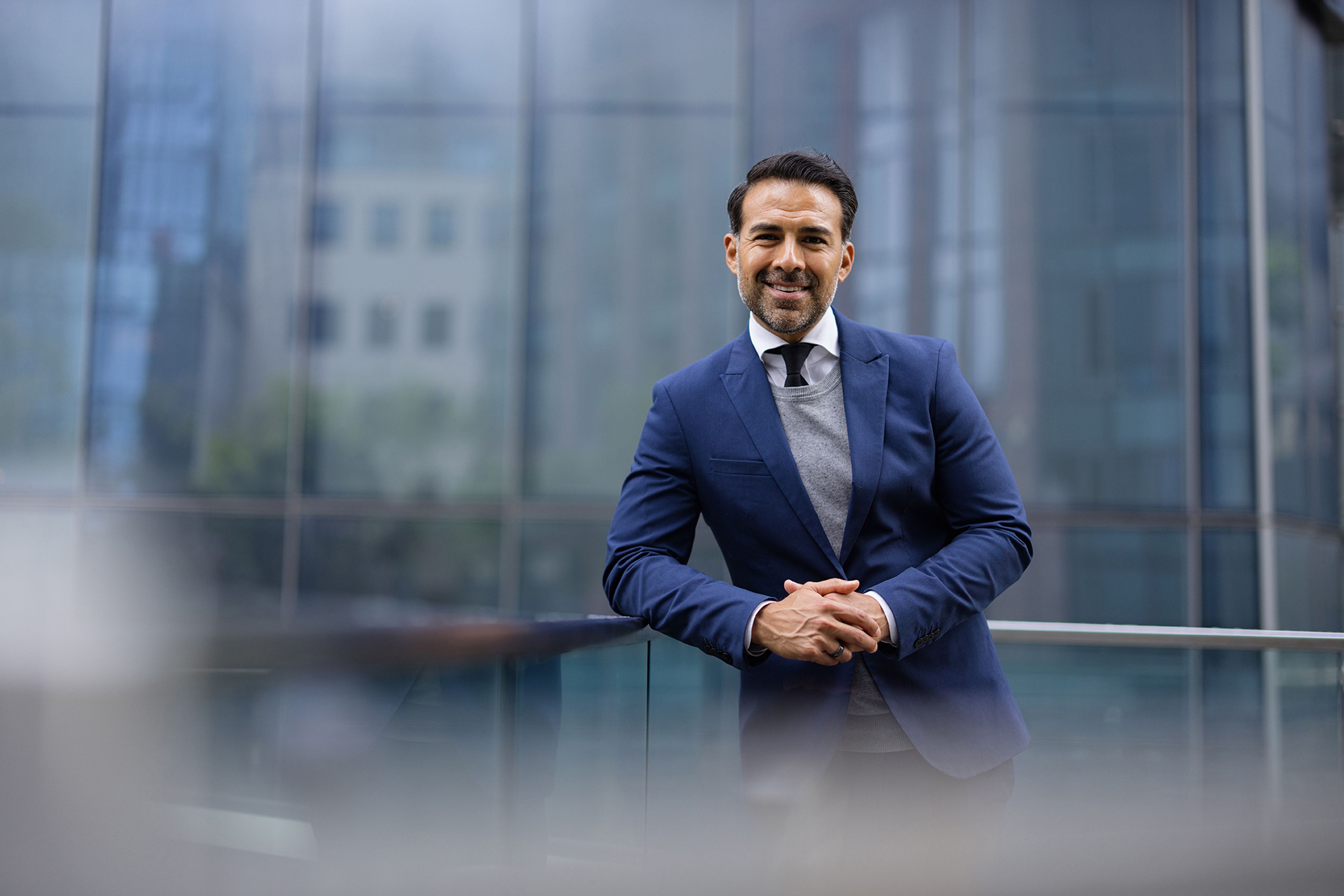 Smiling man in a blue suit leans on a railing, with a modern glass building in the background.