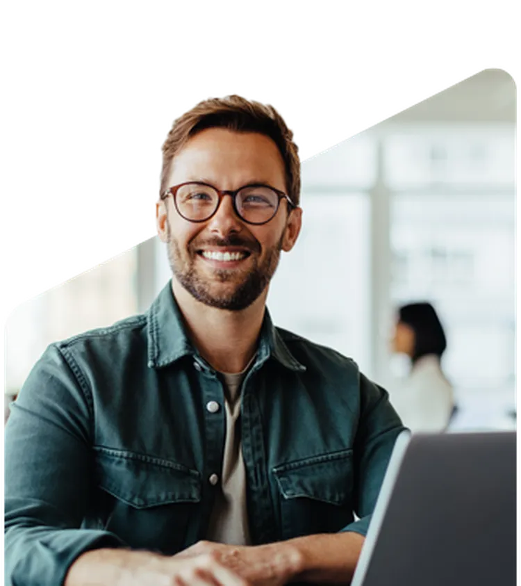 Smiling man with glasses and beard working on a laptop in a bright office, with a blurred colleague in the background.